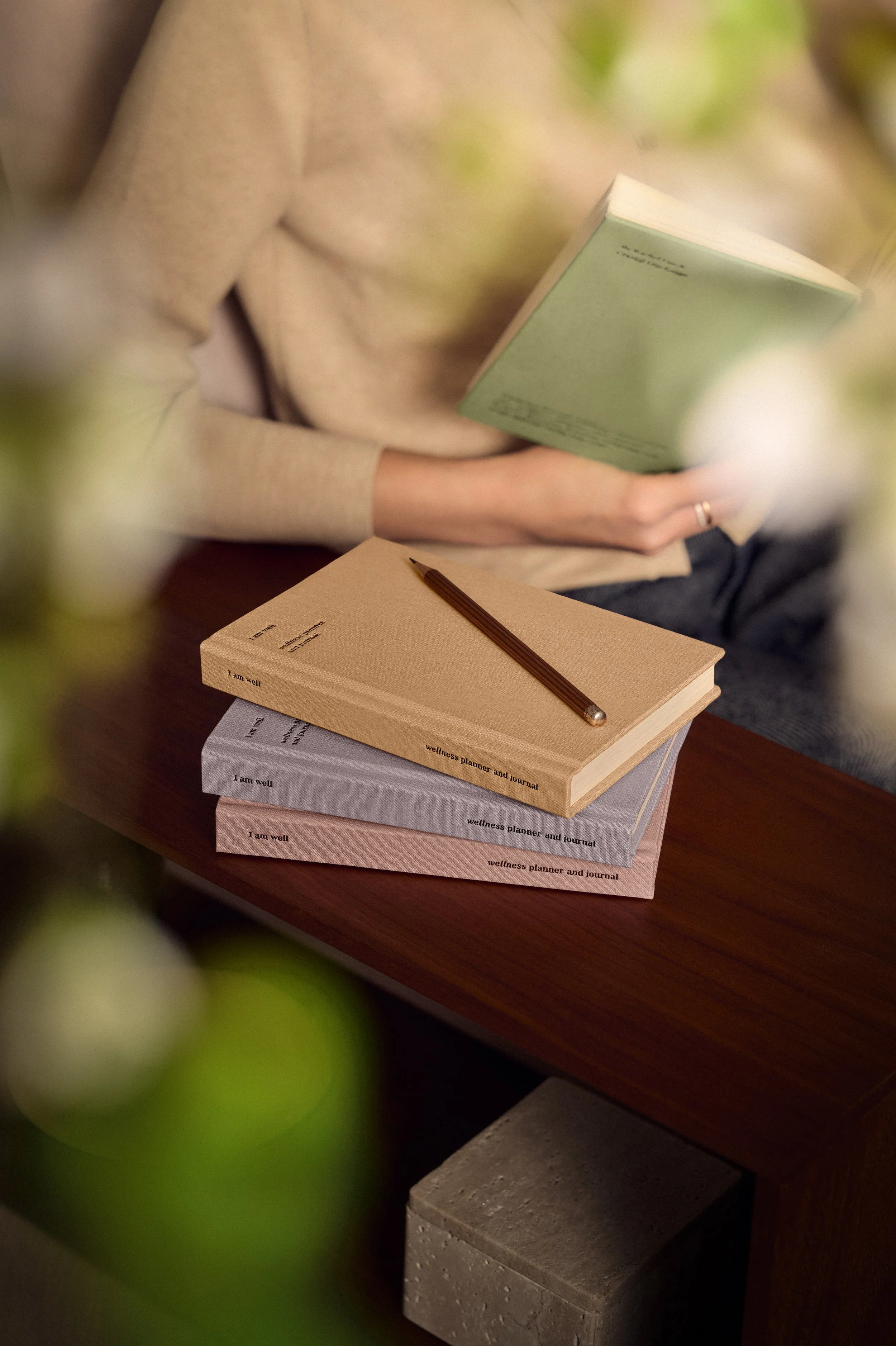 Close-up of a person sitting at a wooden table reading a book, with three notebooks stacked in front of them, one open, and a pencil placed on top of the notebooks.