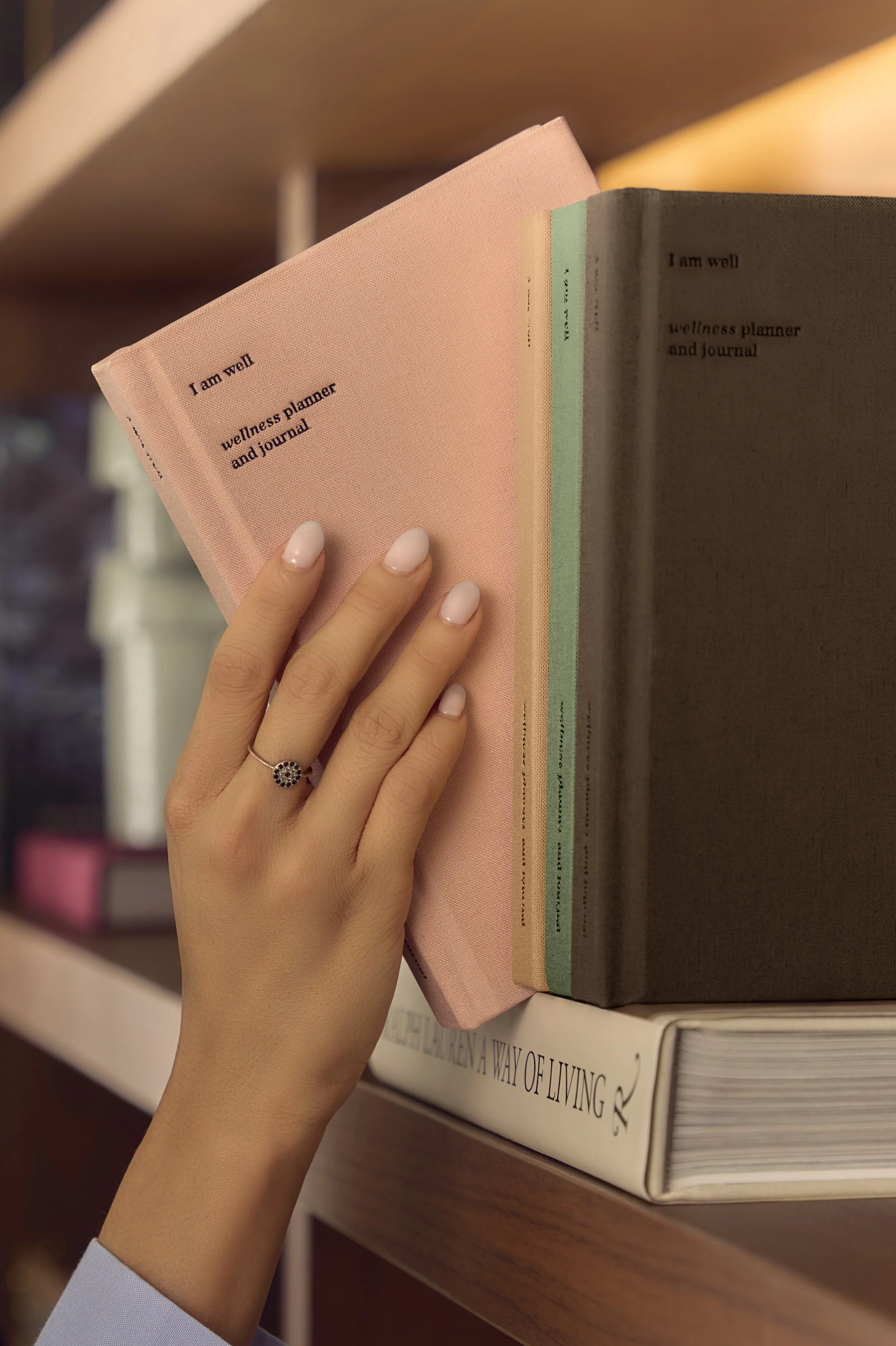 A hand with manicured nails and a ring, holding a pink wellness planner and journal, placed on a shelf with other books.