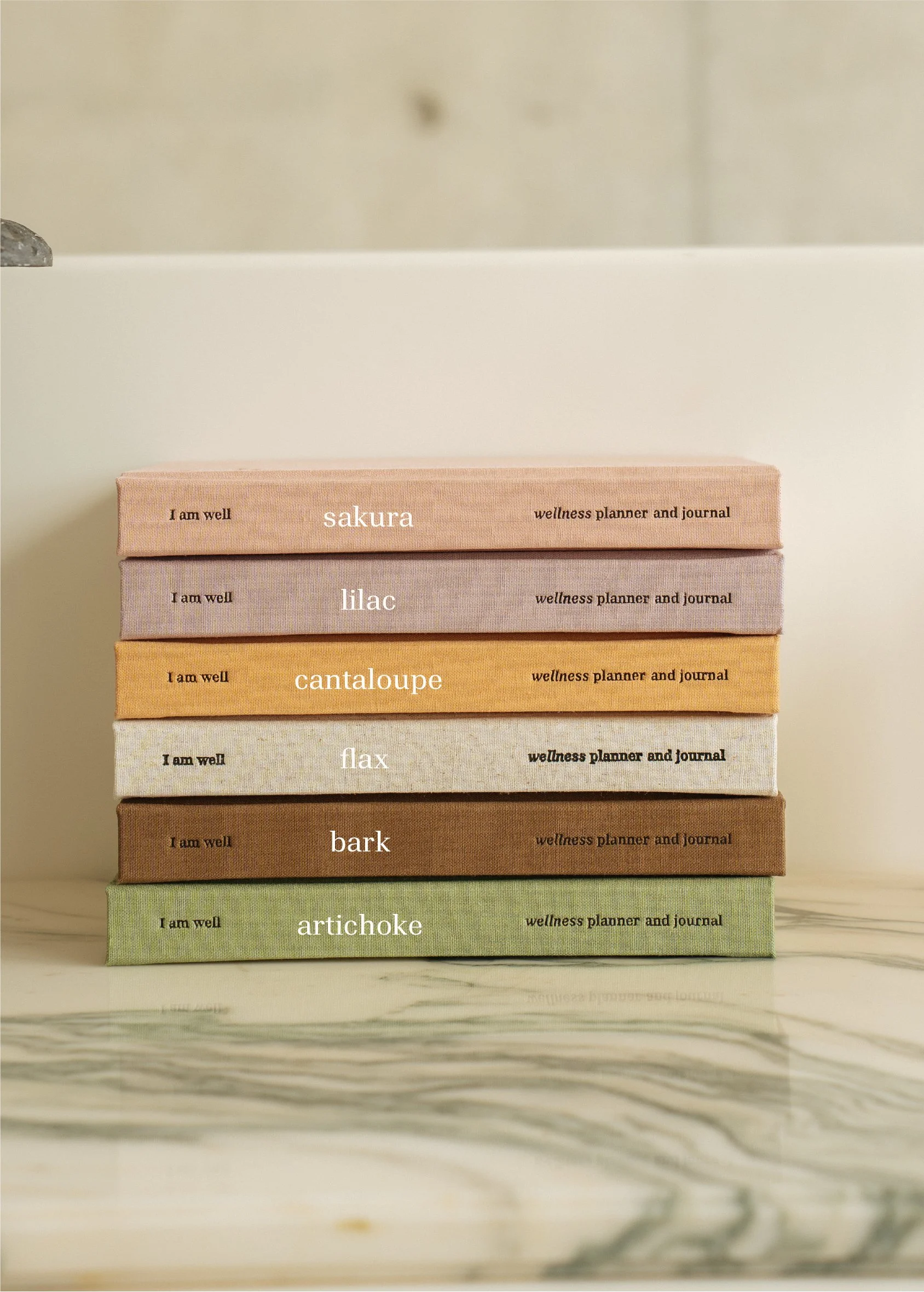 Stack of seven wellness planner and journal books in pastel colors labeled sakura, lilac, cantaloupe, flax, bark, artichoke on a marble surface