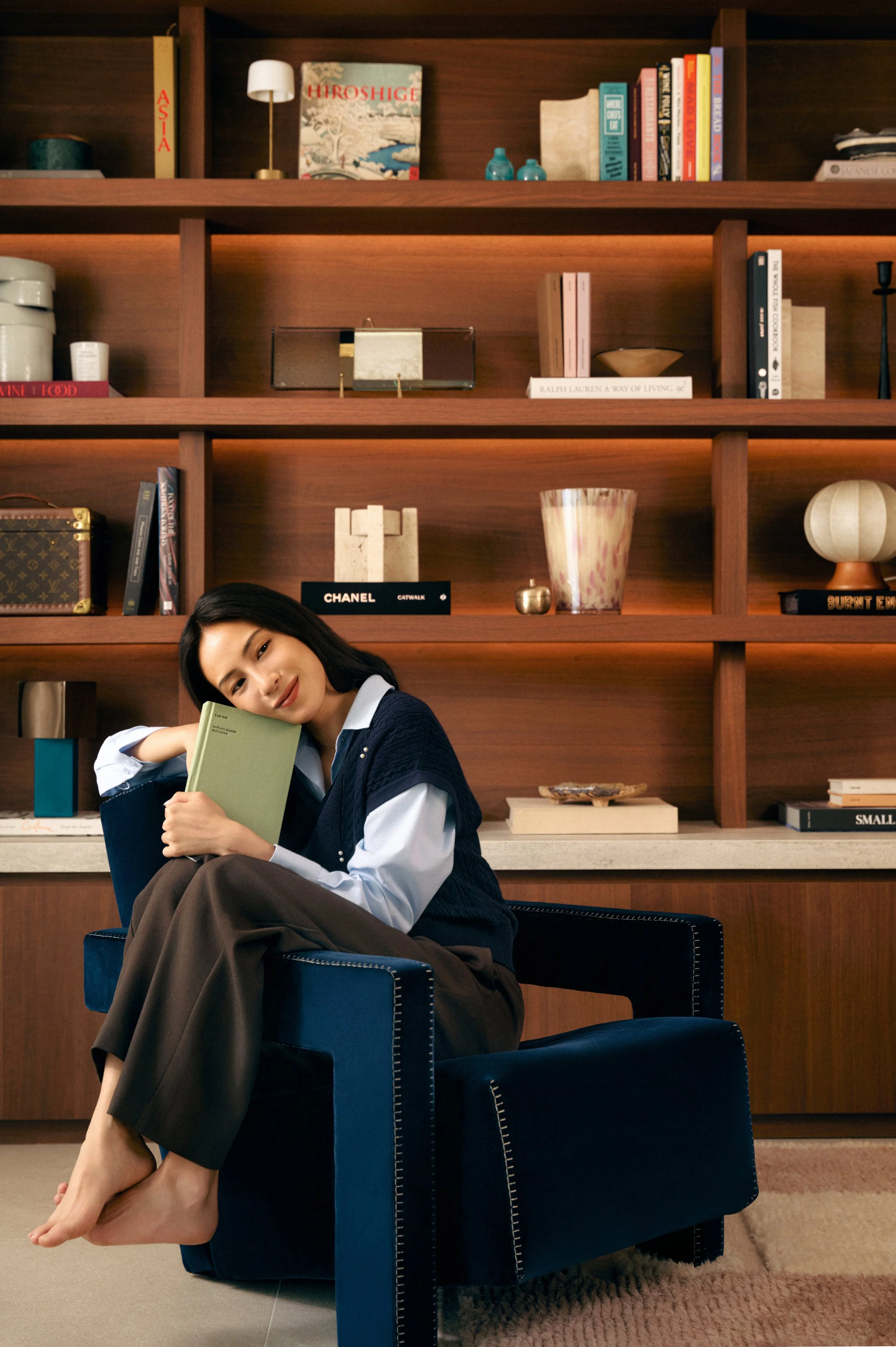 A woman sitting on a navy blue armchair in a room with a wooden bookshelf behind her. She is holding a light green book, resting her head on it, and smiling. She is barefoot, wearing a white shirt, dark blue vest, and brown pants.