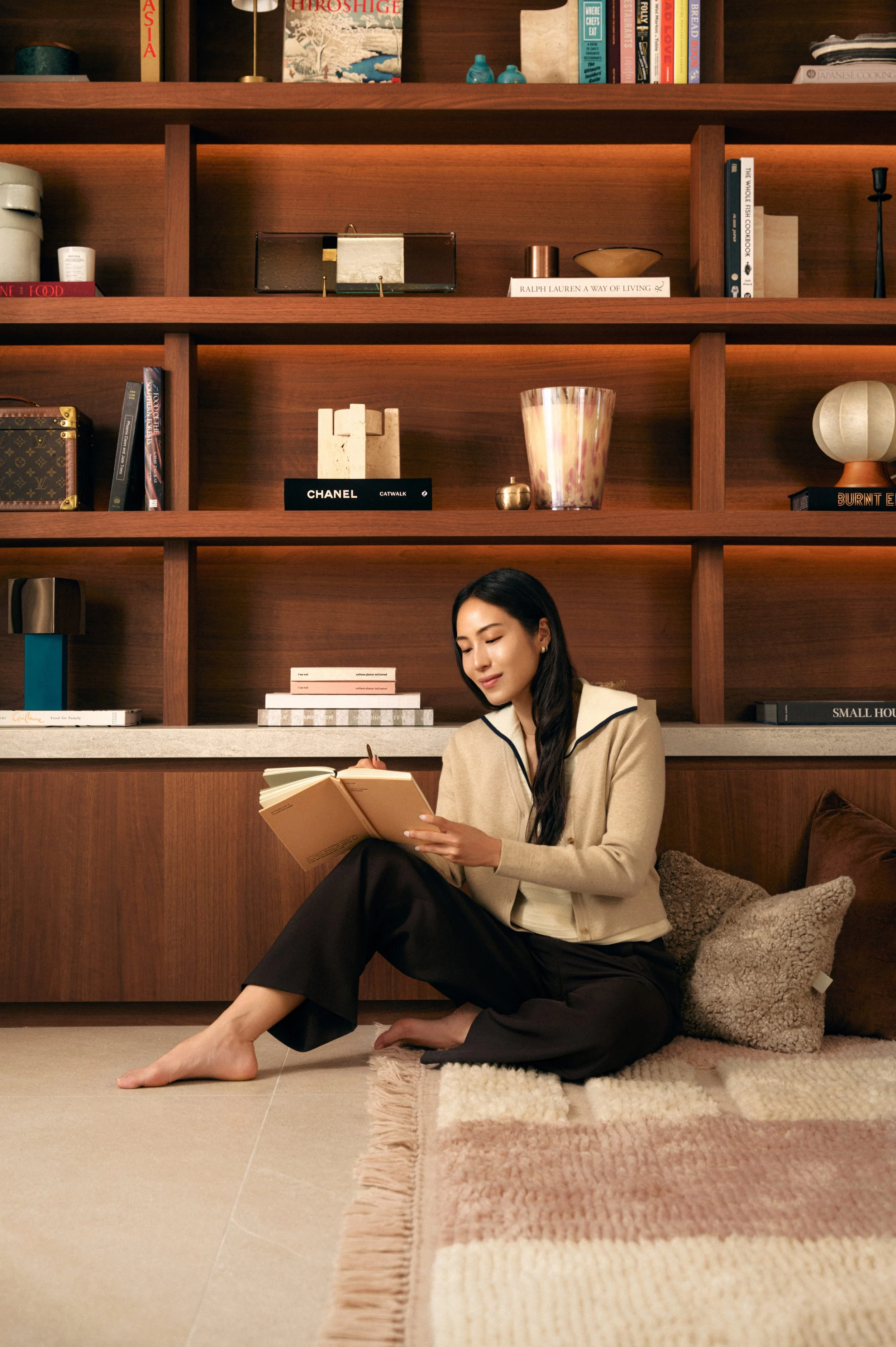 A woman sitting on the floor reading a book in a room with a large wooden bookshelf behind her.