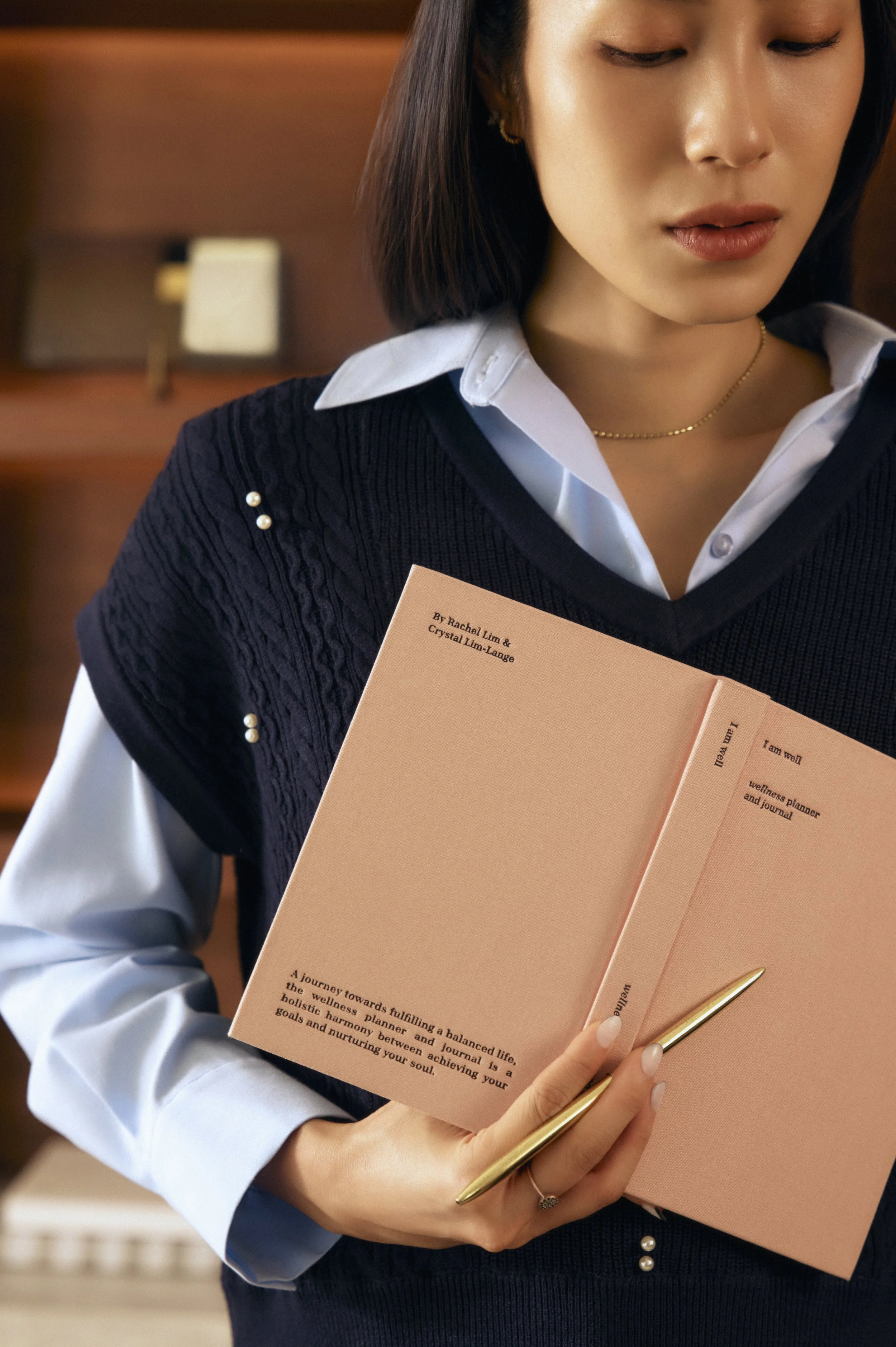 A woman with shoulder-length dark hair wearing a white shirt and black sweater with pearl accents, holding an open pink journal and a gold pen, looking down at the journal.