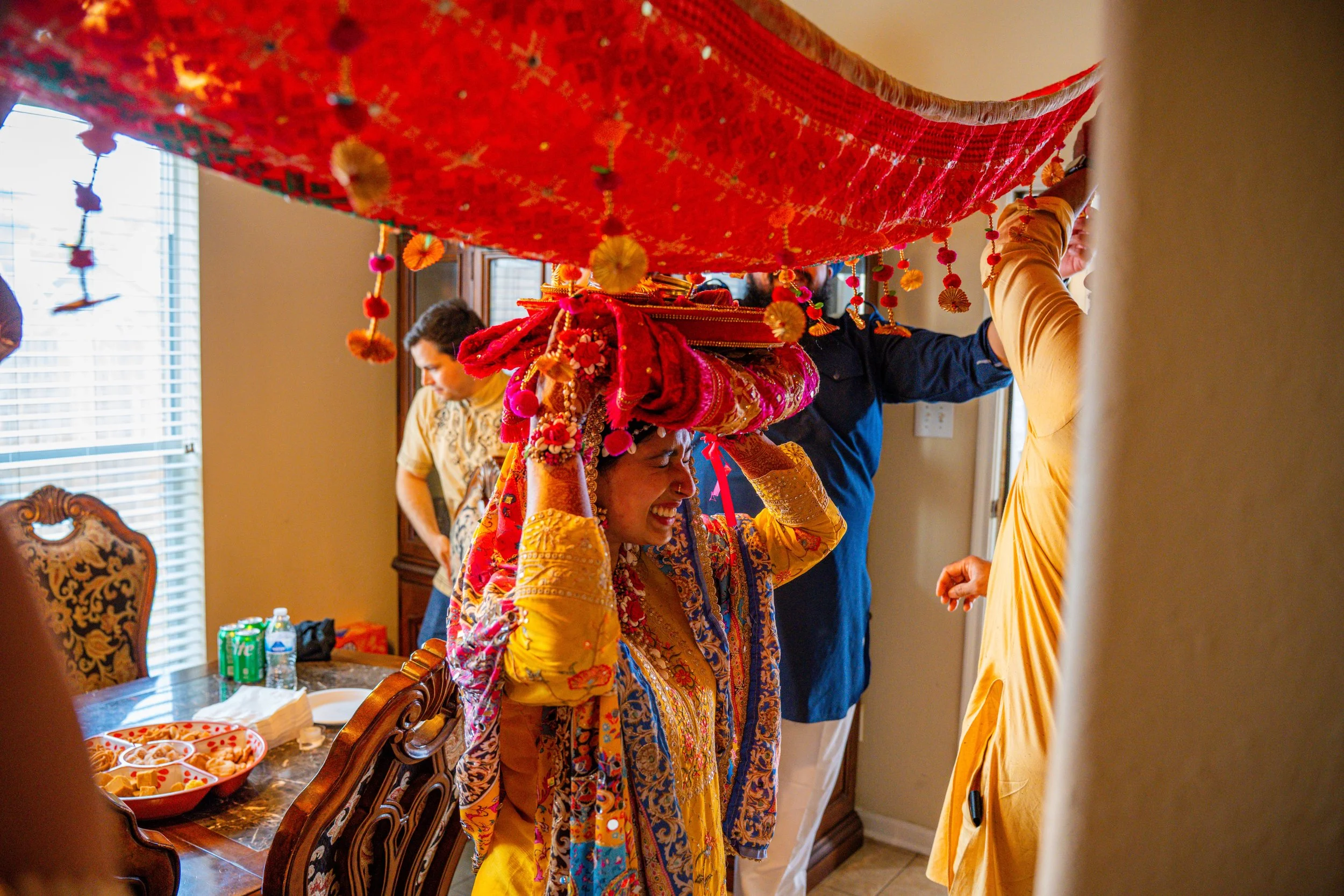 People participating in a traditional Indian wedding ceremony. The bride is smiling and dressed in colorful, ornate attire, carrying a decorated canopy or ornament on her head. The setting appears to be indoors, with a dining table and benches visibl