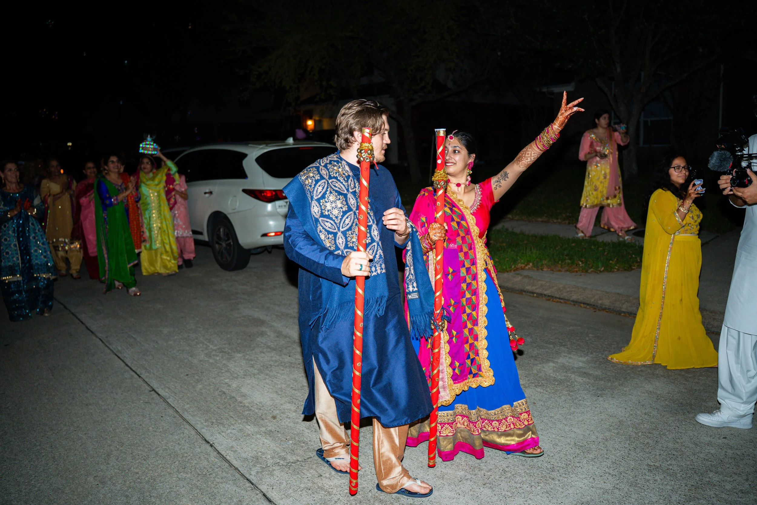 Indian bride and groom in traditional attire walking on street during nighttime, surrounded by friends and family, with a woman gesturing and a man filming, cars parked in background.
