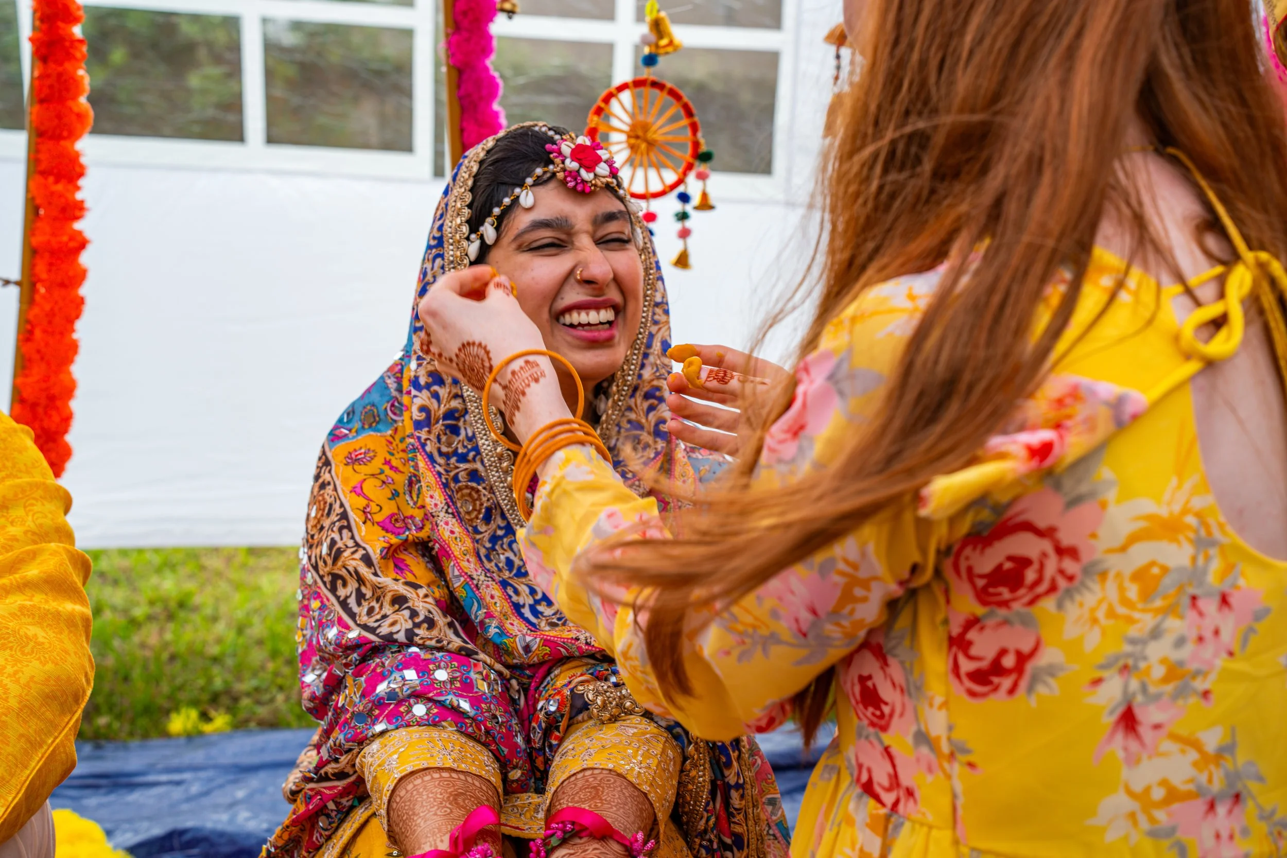 A woman dressed in traditional Indian attire, smiling and applying traditional henna tattoos during a celebration or ceremony, with colorful decorations in the background.