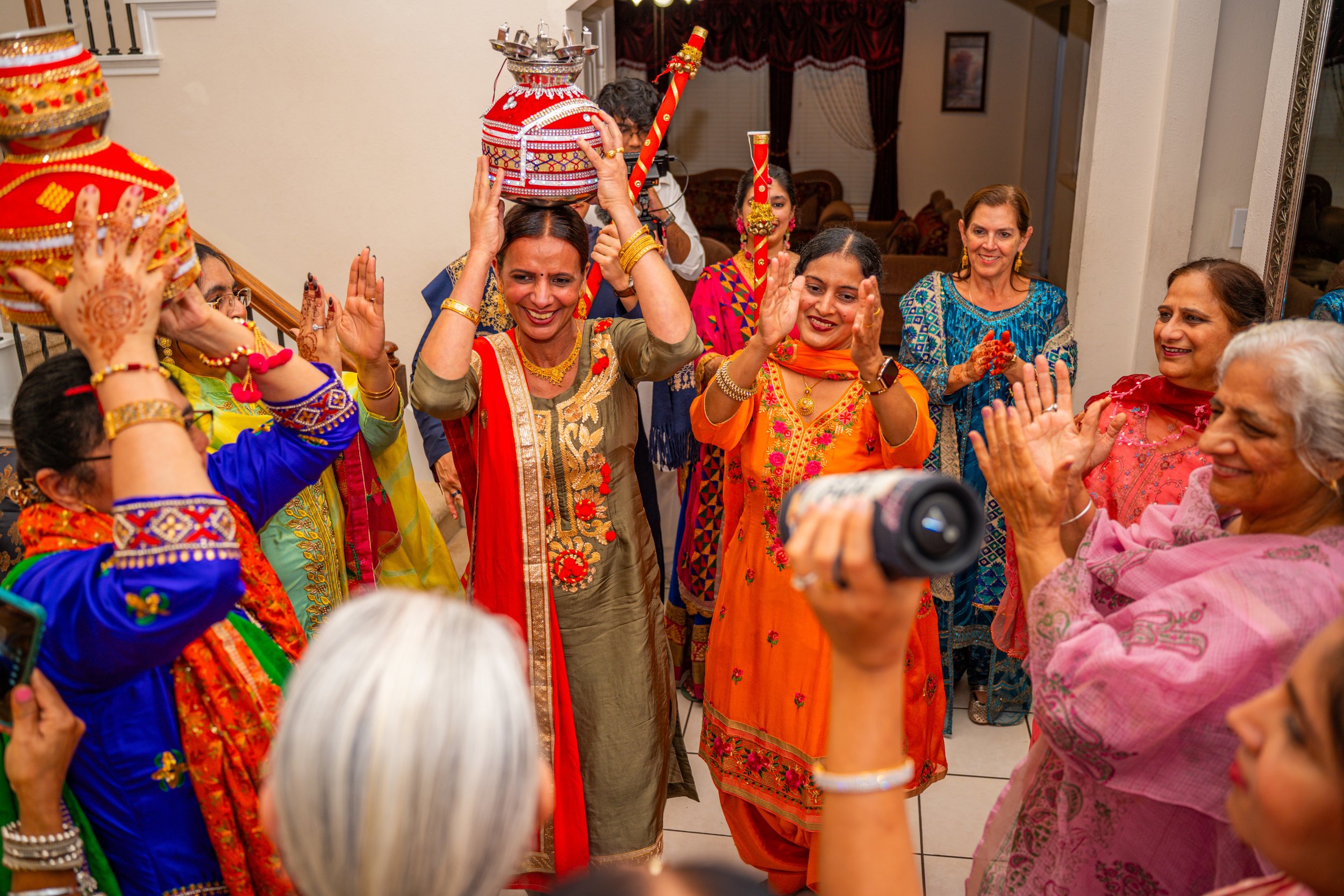 A group of women in colorful traditional Indian sarees and kurtas celebrating a wedding or festival indoors. They are wearing jewelry and headpieces, clapping, smiling, and dancing. One woman is holding a decorated pot on her head.