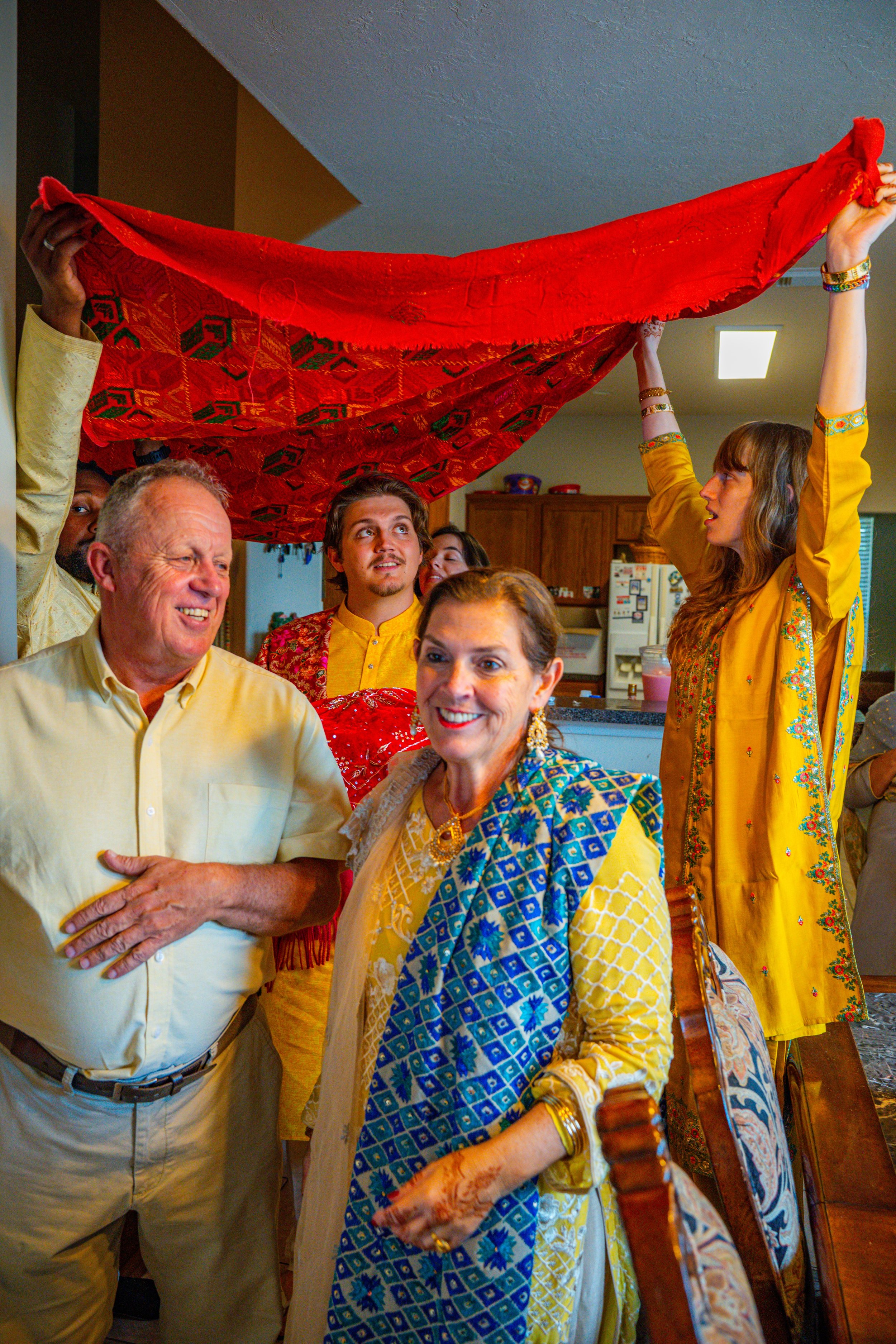 A group of people celebrating an Indian or South Asian cultural event, with women wearing traditional colorful sarees and men in casual attire. Two women are holding a bright red cloth or shawl above the group.