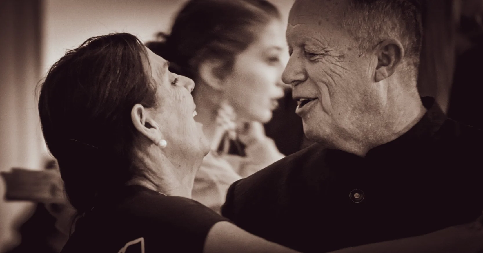 An elderly man and woman are dancing closely together, smiling and gazing into each other's eyes. In the background, there are two women, one with her eyes closed and another with her eyes open, both appearing to be enjoying the moment.