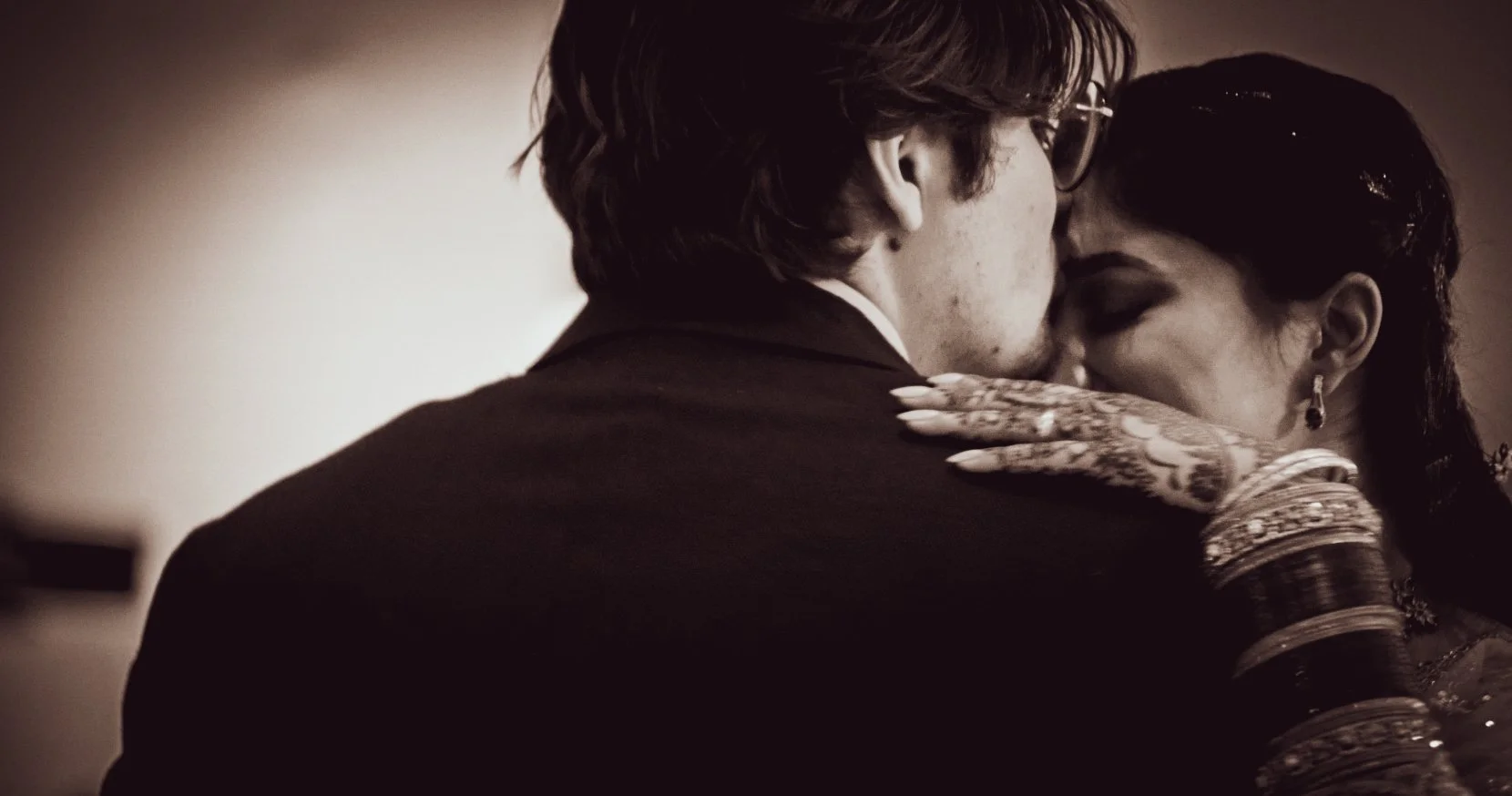 A close-up, black-and-white photo of a man kissing a woman on the forehead, with the woman's eyes closed and her hand resting on the man's shoulder. The woman is wearing traditional jewelry and bangles, and has henna on her hand.