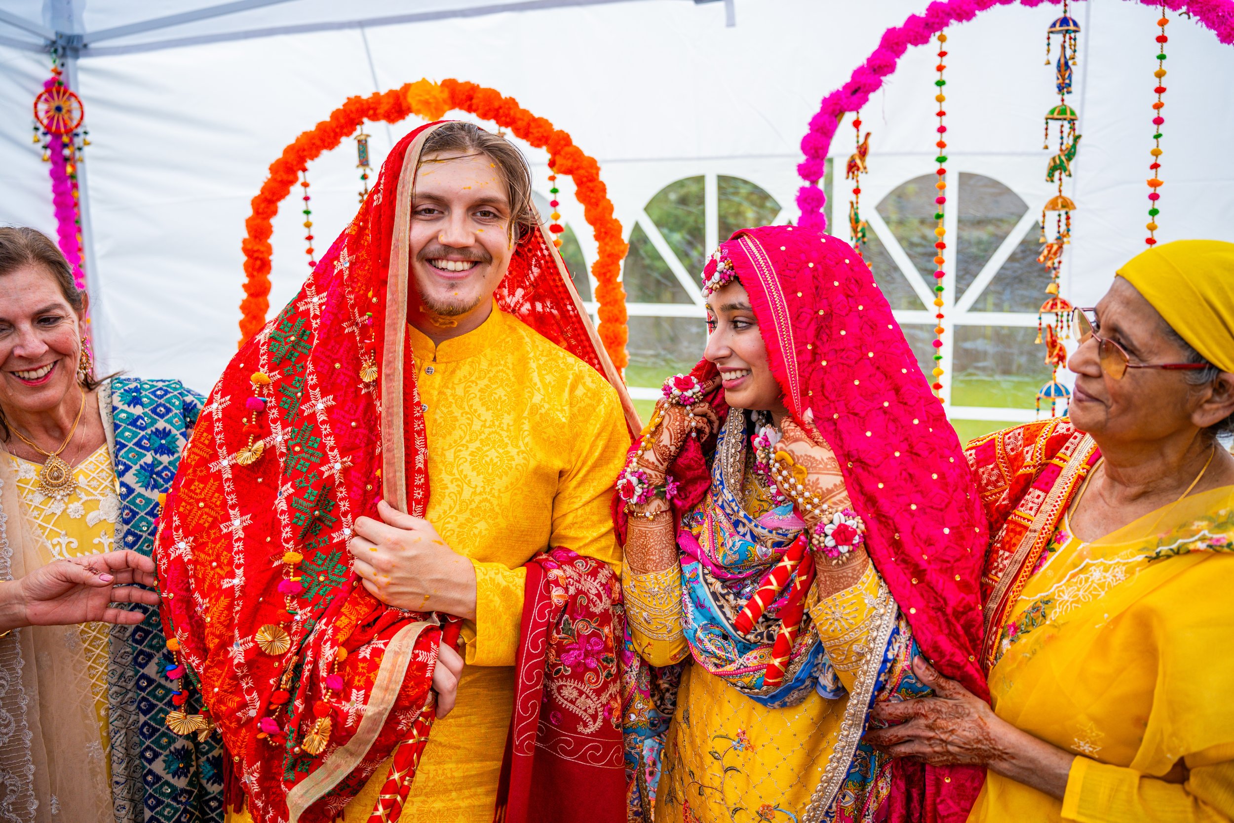Wedding ceremony with the groom and bride dressed in traditional Indian attire, surrounded by family members in colorful clothing with floral decoration in the background.