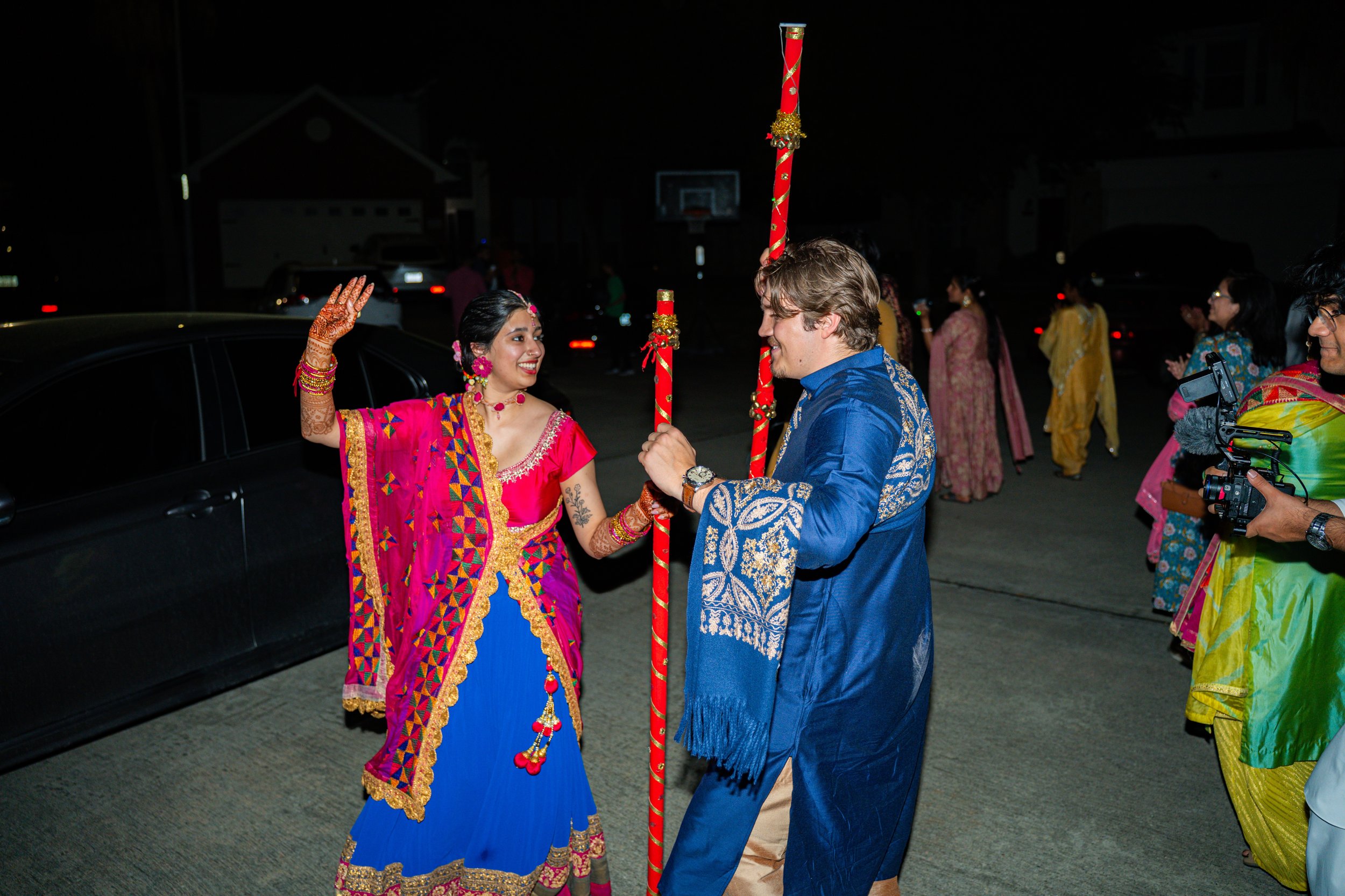 People celebrating during a nighttime Indian wedding, with a woman in a pink and blue traditional dress dancing and a man in a blue kurta holding a decorated staff, surrounded by guests in colorful attire.
