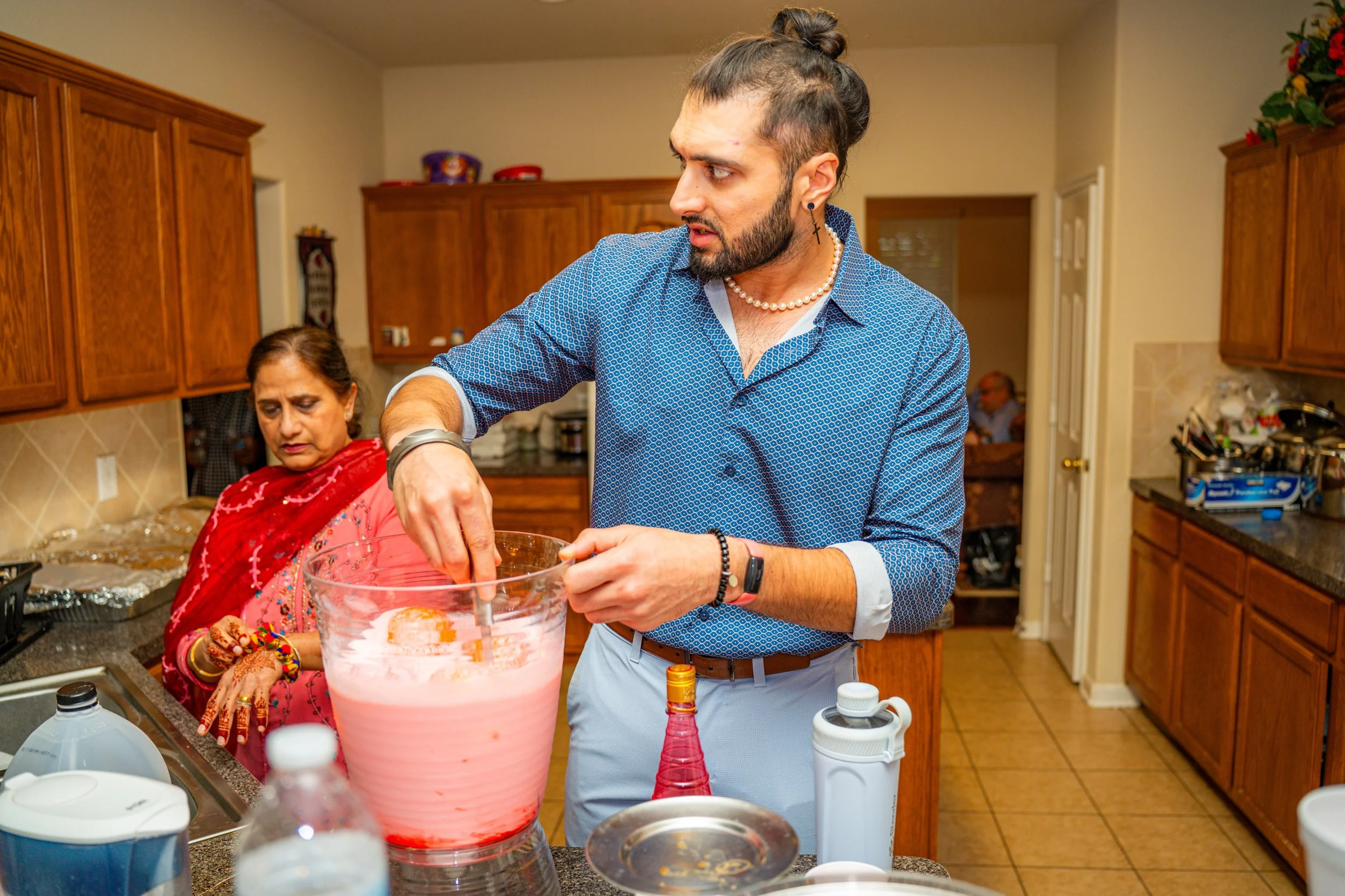 A man with a beard, tie-dye shirt, pearl necklace, and earrings making a drink in a glass bowl in a kitchen, with an older woman in traditional Indian attire and jewelry in the background.