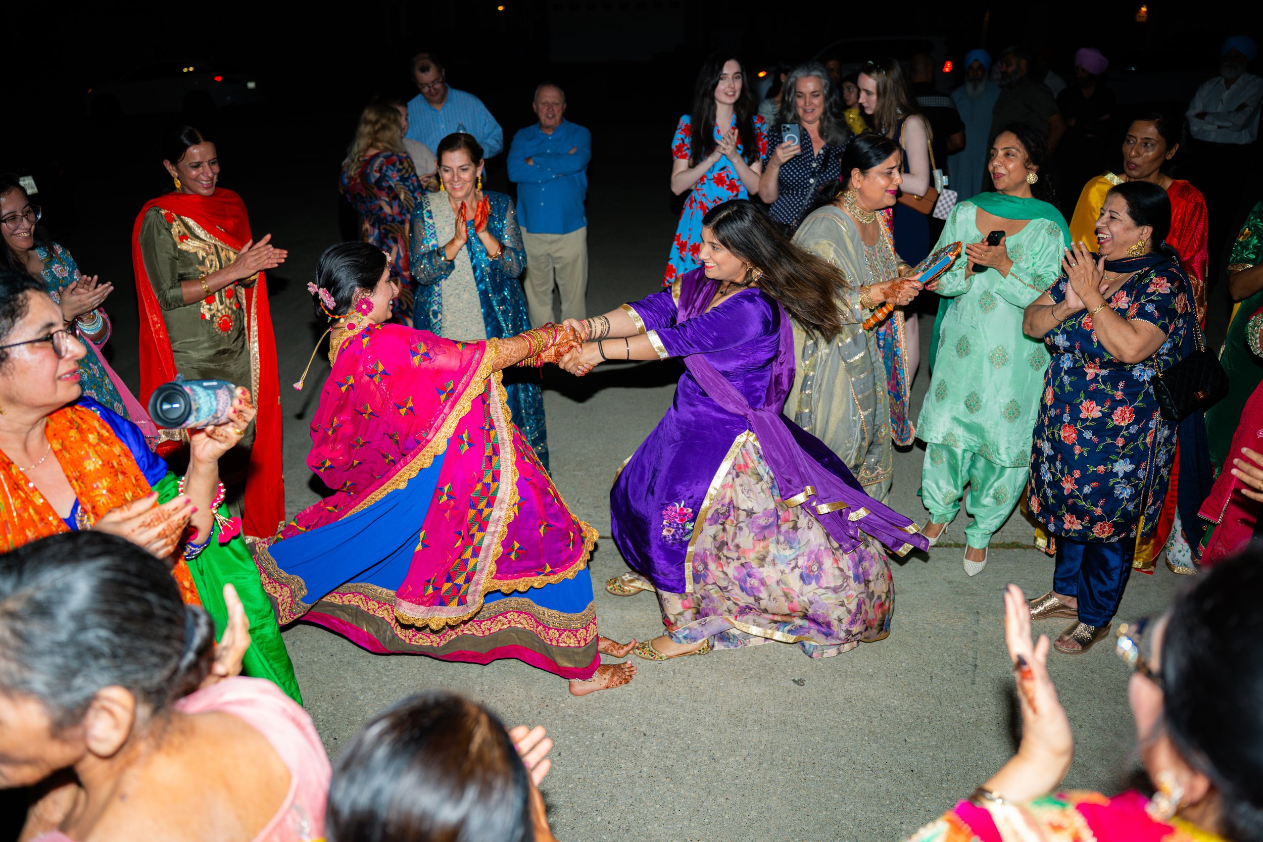 A group of women dancing and celebrating at an outdoor event during the night, dressed in colorful traditional Indian clothing, with some clapping and smiling.