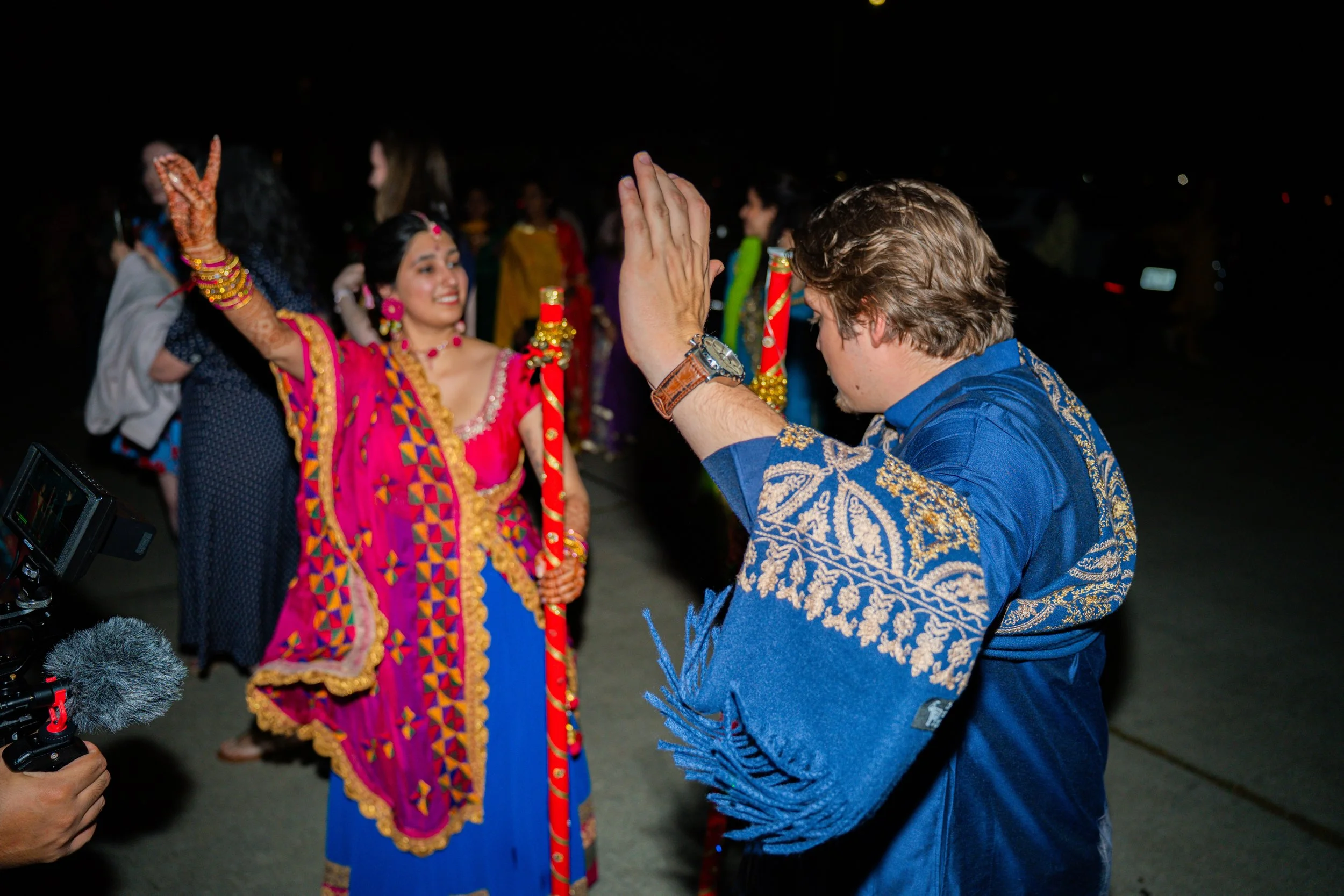 People celebrating at a nighttime event, with a woman in colorful traditional Indian attire holding candles and a man in a blue embroidered shirt joining the celebration.