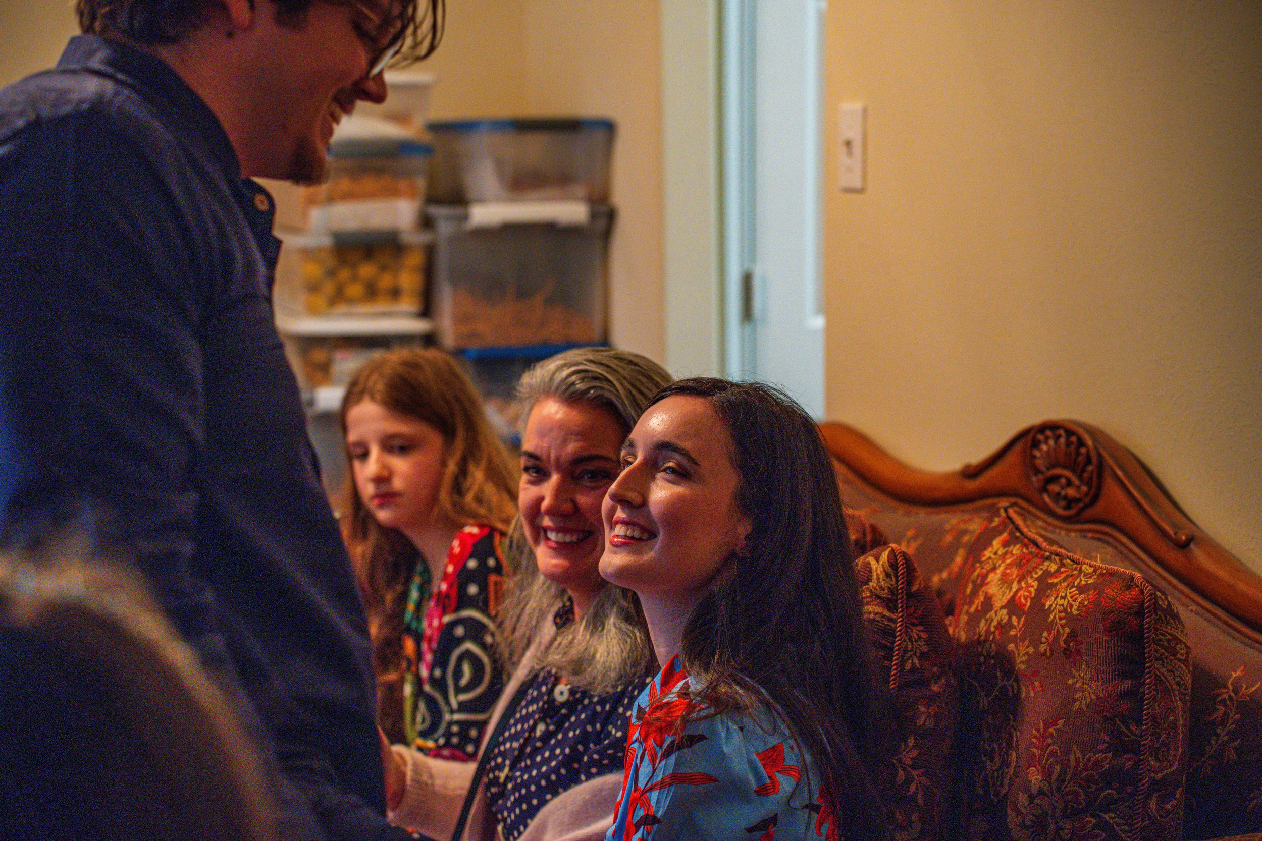 Four people sitting on a patterned couch, smiling and engaging in conversation with a standing person in front of them, in a cozy room with shelves of storage containers in the background.