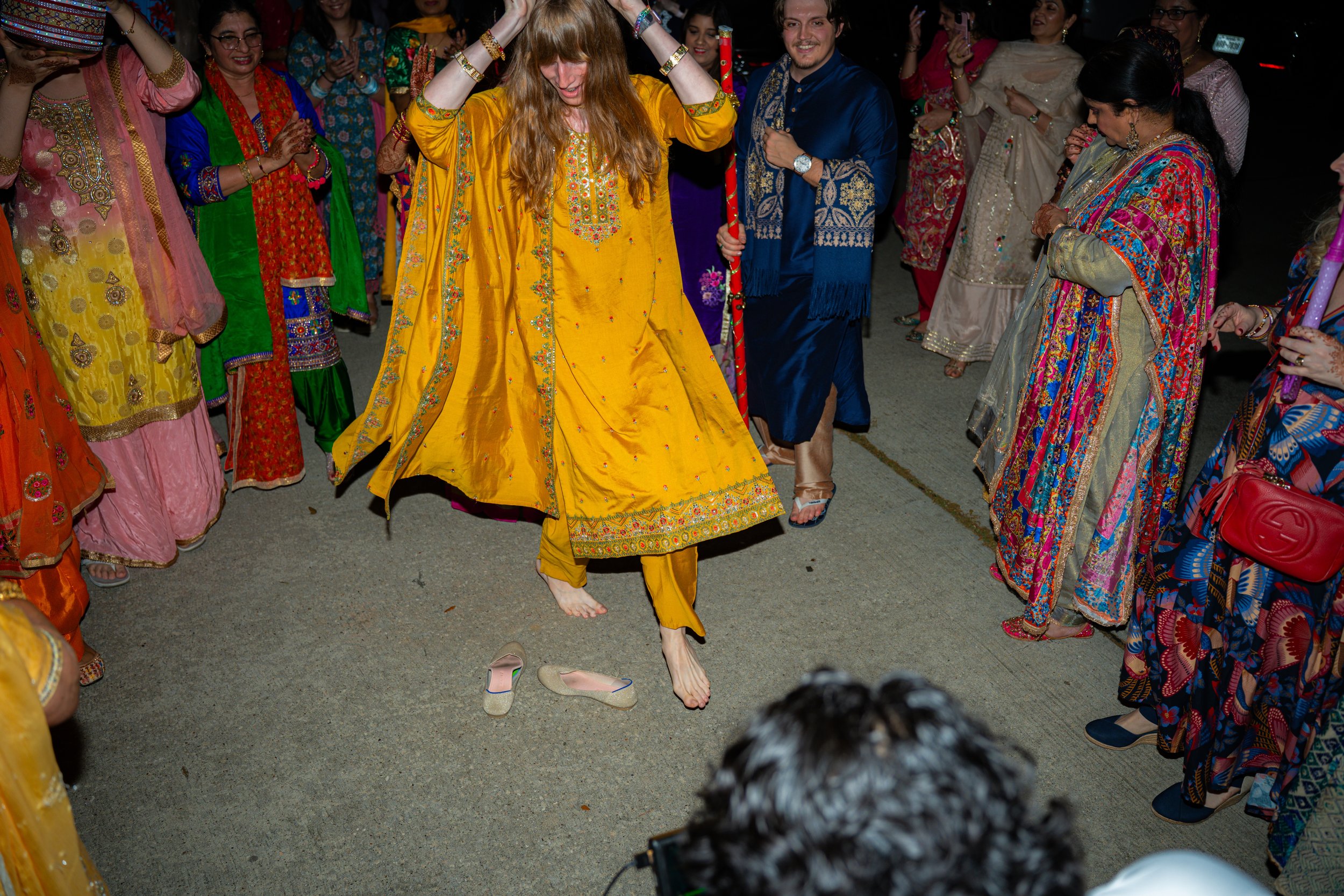 A woman in a bright yellow traditional dress is dancing barefoot at an event, surrounded by people dressed in colorful traditional attire, some clapping and holding hands.