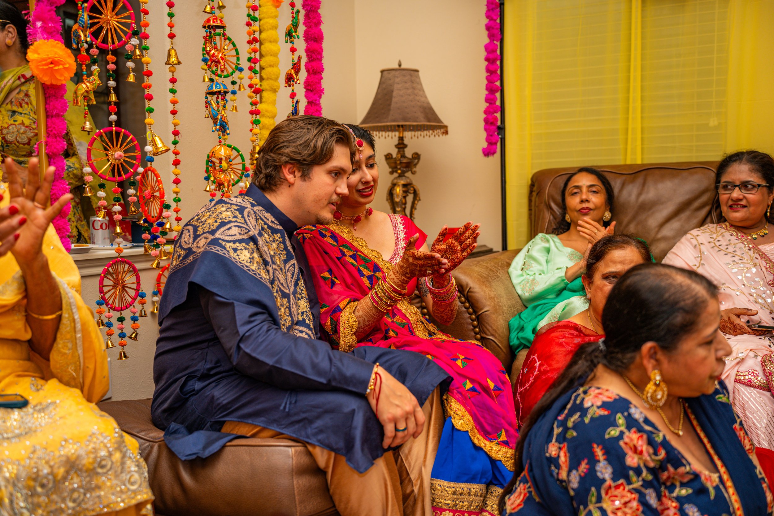 Group of people dressed in colorful traditional Indian attire, sitting on a sofa and engaging in a celebration.