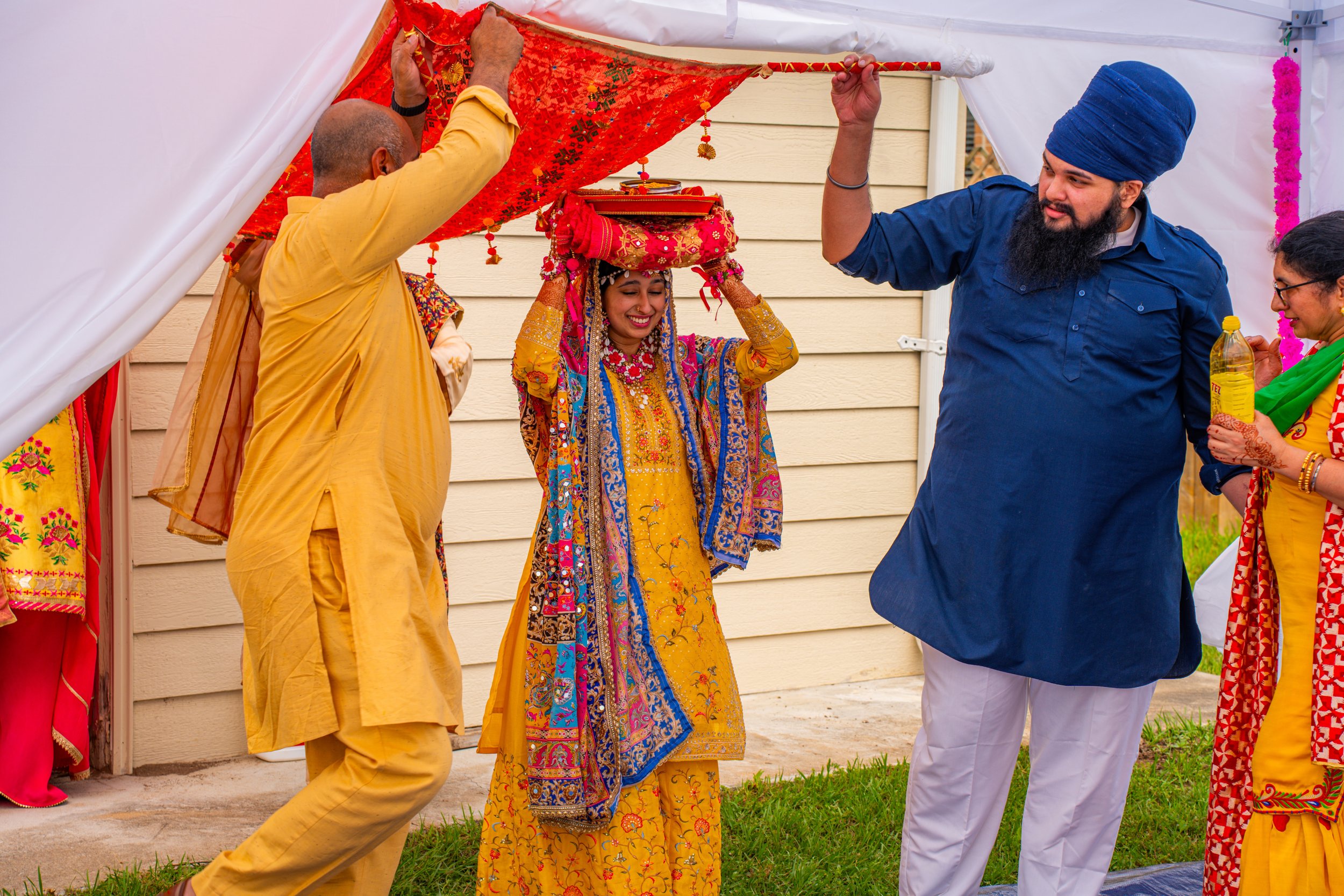 Indian bride in traditional colorful wedding attire smiling as she walks under a decorated canopy, surrounded by family members during a wedding ceremony.