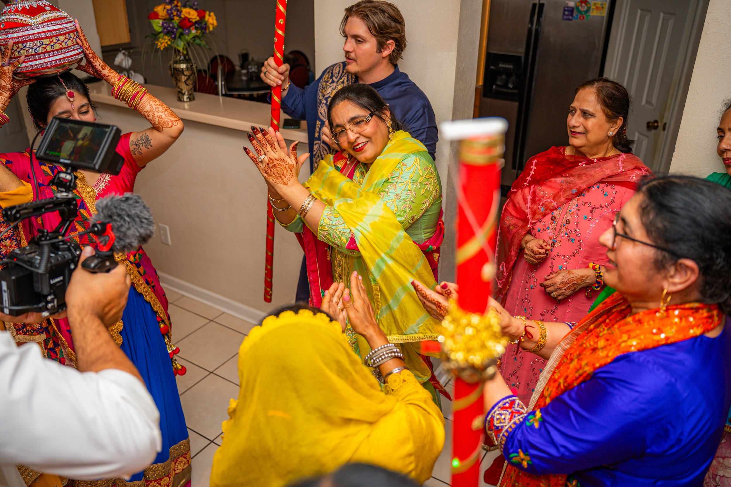 Group of women in colorful traditional Indian attire participating in a celebration inside a home, with decorated items and a camera capturing the moment.