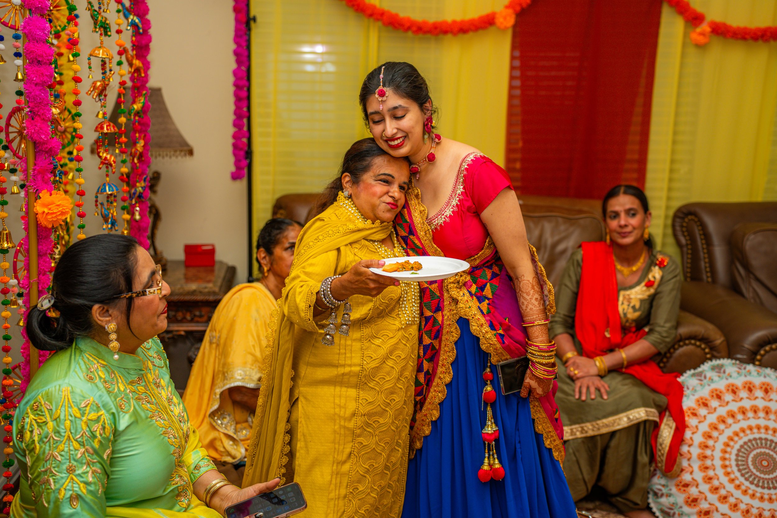 Women wearing colorful traditional Indian attire at a celebration with vibrant decorations, including hanging flowers and fabric drapes.