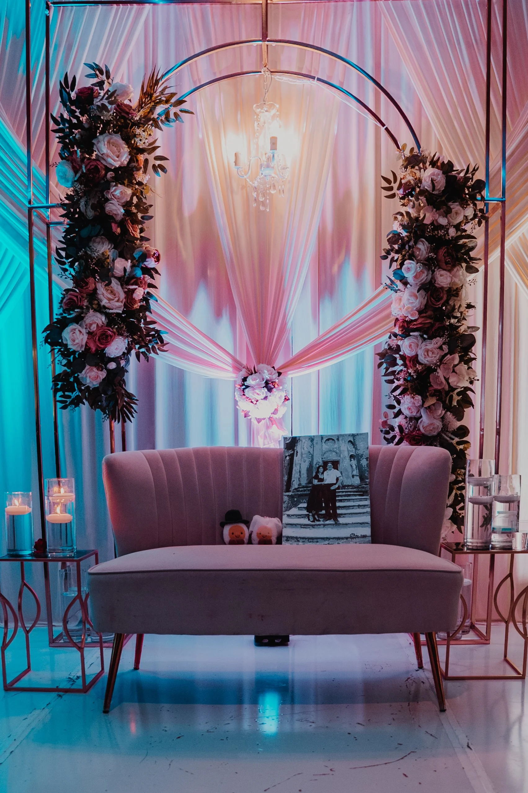 A decorated backdrop with pink, white, and purple flowers, draped pink fabric, and a chandelier above a pink vintage-style sofa. There are small tables with candles and plush toys, and a framed black-and-white photo on the sofa.
