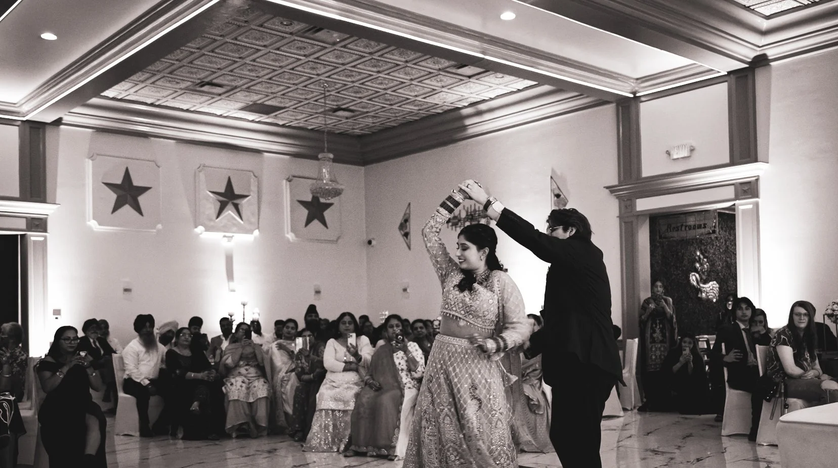 A couple dancing in traditional attire surrounded by seated guests in a decorated hall.
