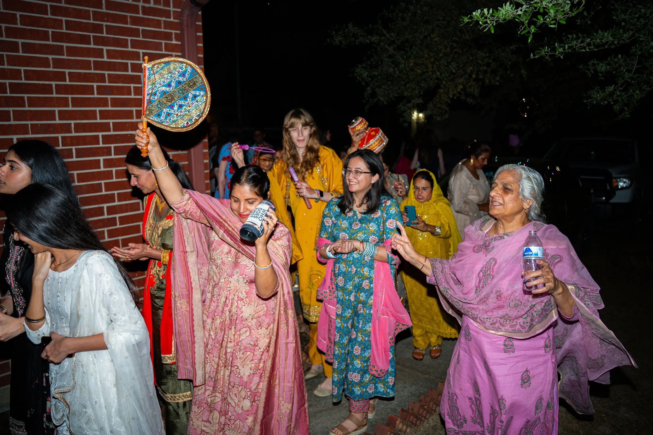 A diverse group of people celebrating at an outdoor event during the night, dressed in colorful traditional Indian attire, some dancing and holding items like a water bottle, a fan, and a small drum.