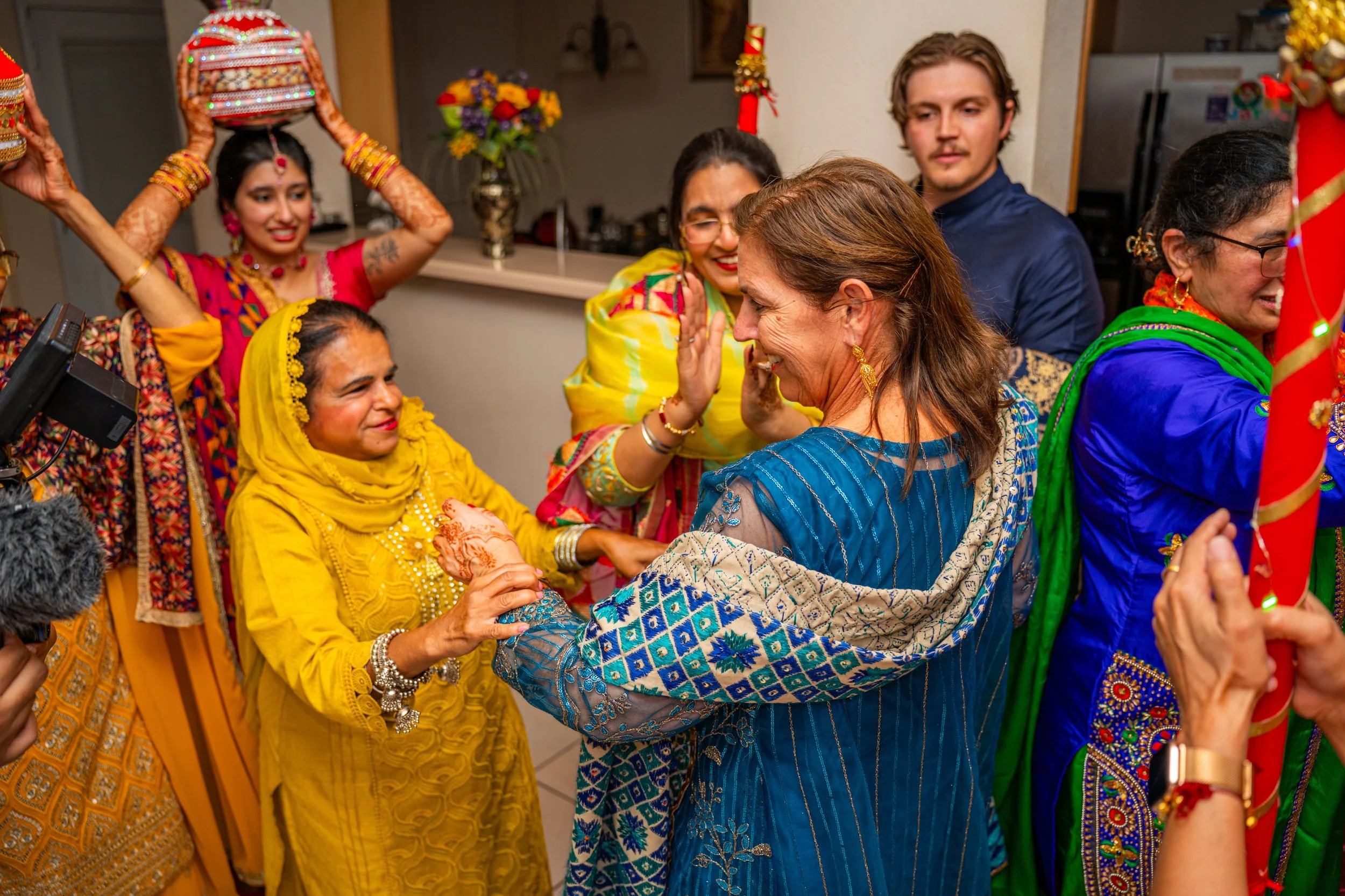 A group of women dressed in traditional Indian attire celebrating a joyous occasion, with some women smiling, clapping, and engaging in a ritual, in a room decorated with flowers and festive decorations.