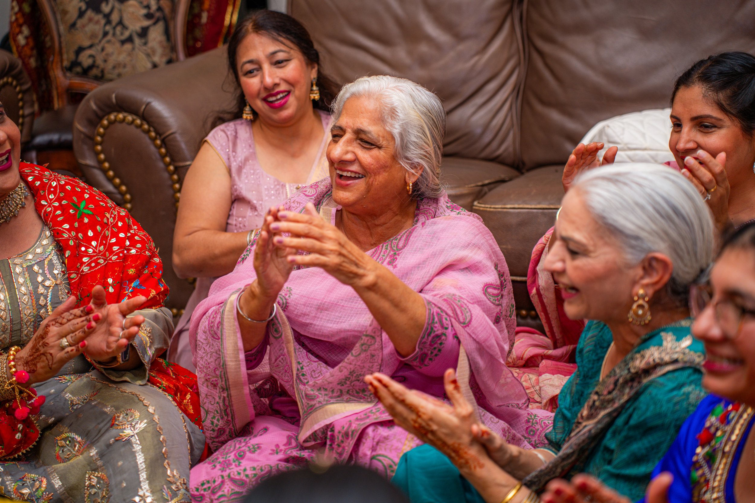 Group of women, mostly elderly, gathered indoors, laughing and smiling, dressed in colorful traditional Indian clothing, sitting on a couch and engaging with each other.