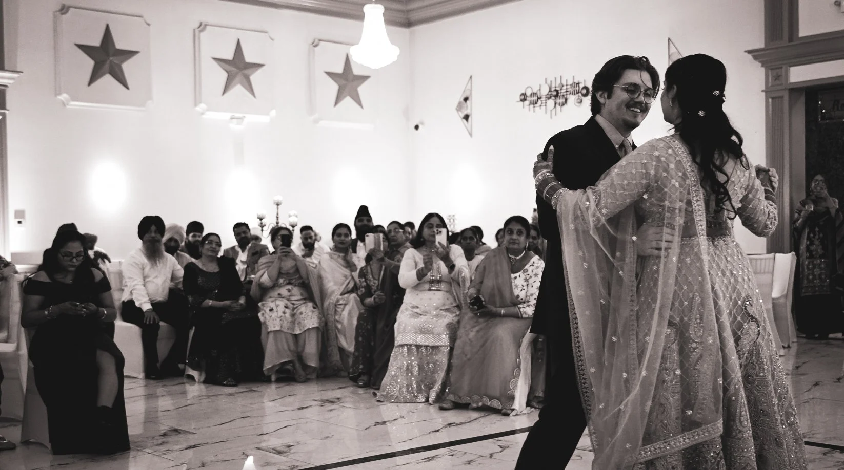 A black and white photo of a couple dancing at a celebration with an audience watching, some taking photos. The room is decorated with stars on the wall and elegant lighting.