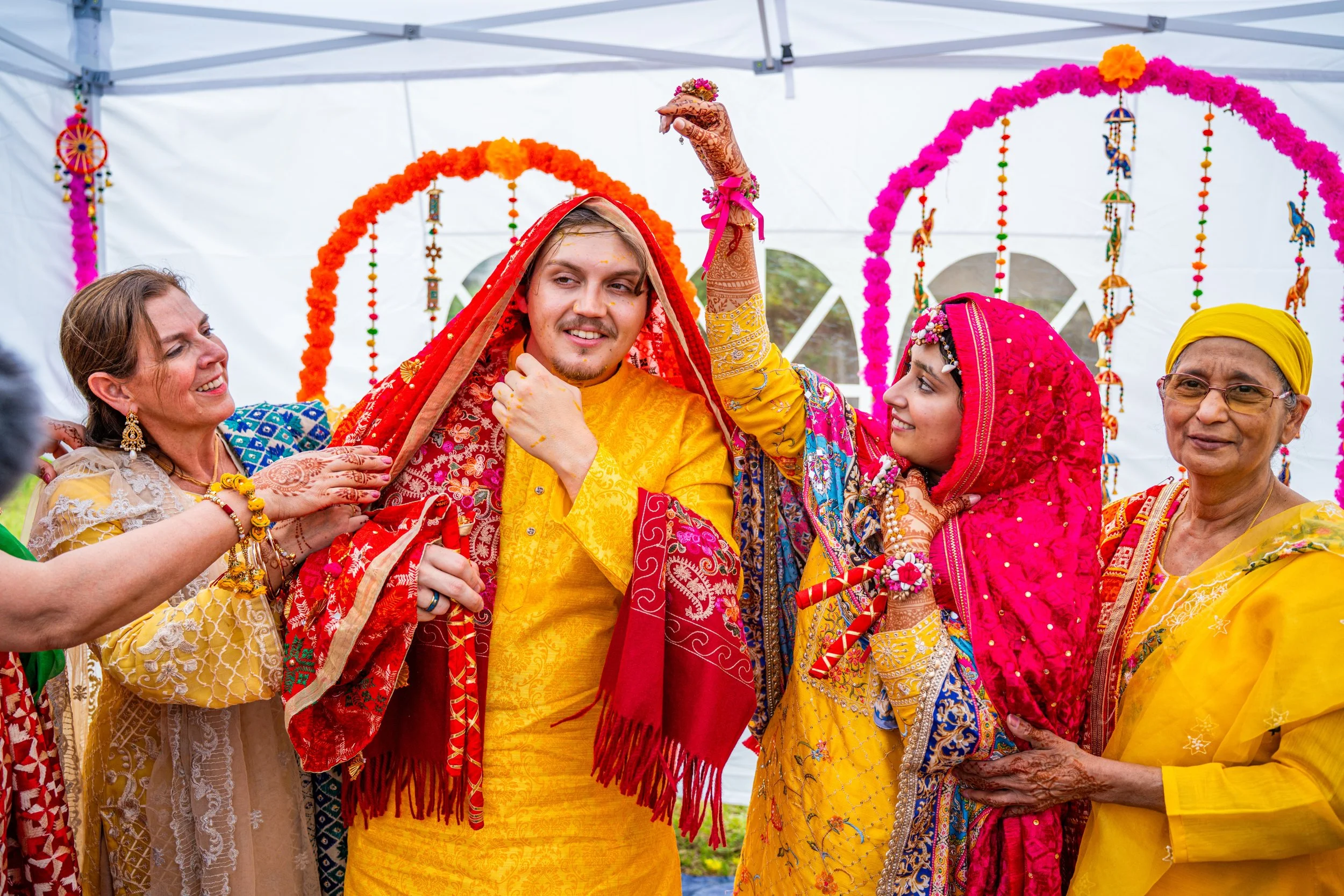 A traditional Indian wedding ceremony with the groom and bride dressed in colorful attire, surrounded by family members under decorative flower arches.