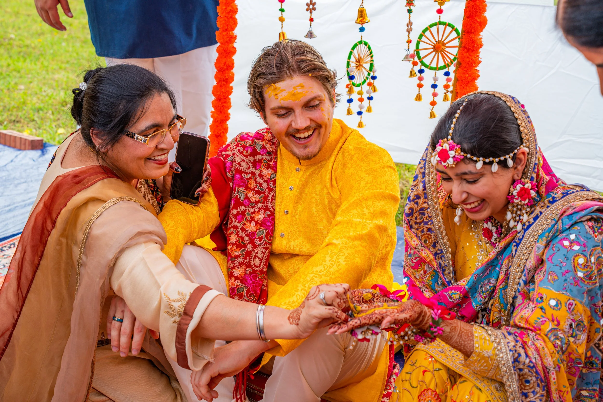 A group of people wearing traditional Indian attire celebrating a joyful event outdoors. They are smiling and engaging in a cultural ritual, decorated with colorful ornaments and flowers.