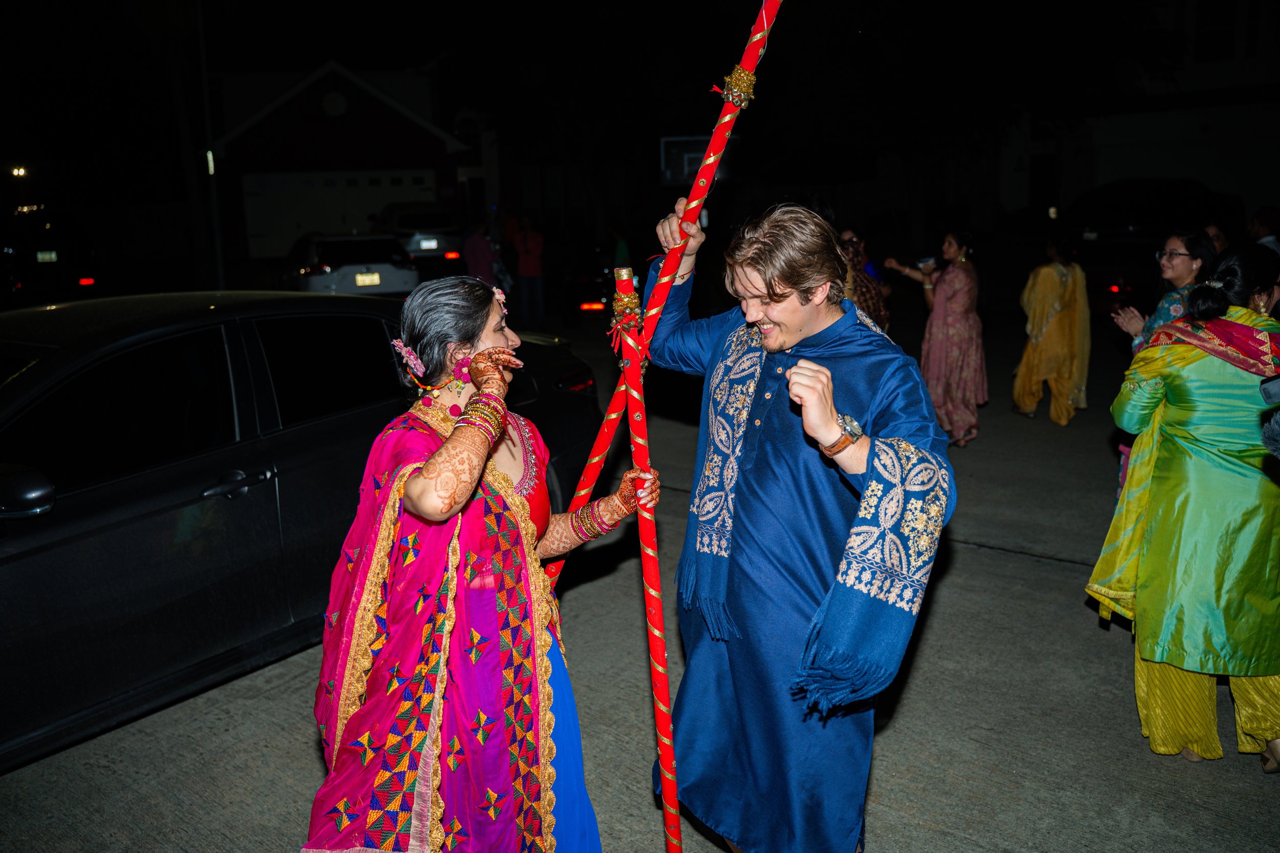A man and woman dressed in traditional Indian wedding attire exchanging joyful expressions during a celebration at night. The woman is wearing a pink and gold saree with intricate henna designs on her hand. The man is wearing a blue sherwani with gol