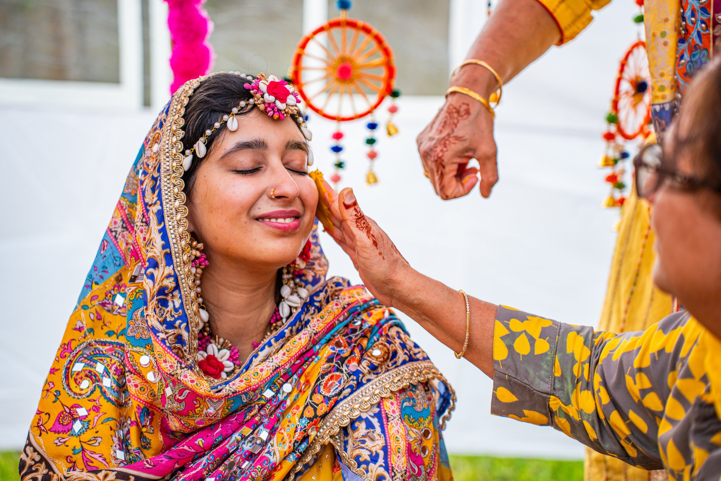 A woman in traditional South Asian attire participating in a wedding ceremony, with a woman applying turmeric on her face.