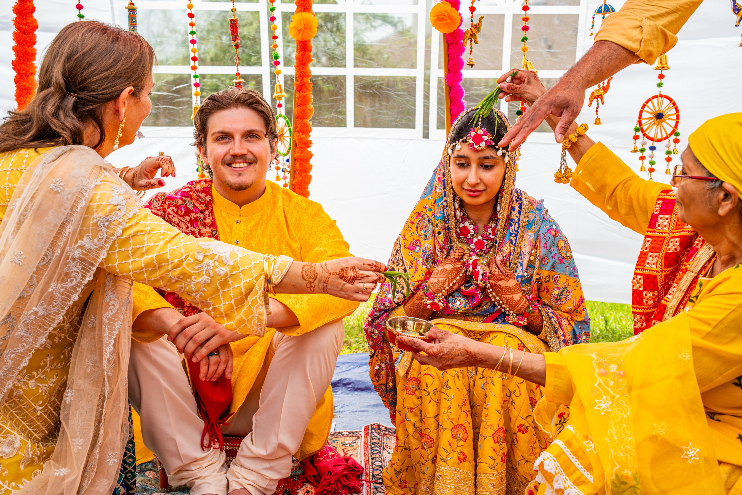 Indian wedding ceremony with a bride and groom seated, surrounded by family performing rituals under decorated canopy.