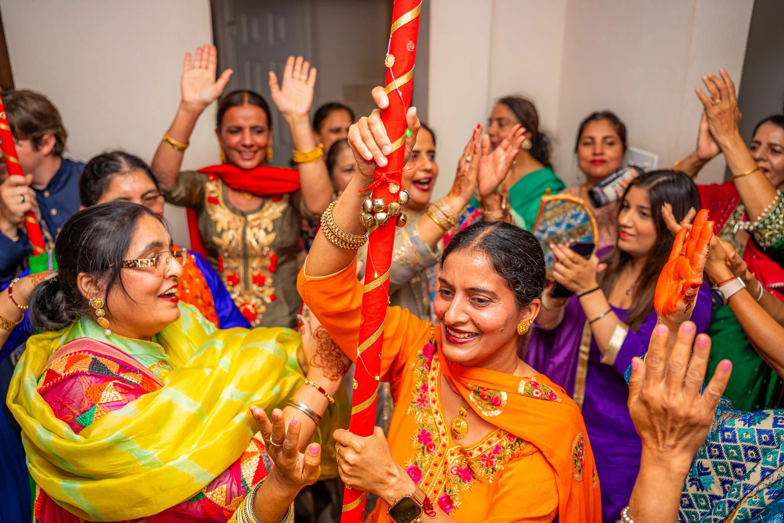 Group of women dressed in colorful traditional Indian attire celebrating together, some with henna on their hands, smiling and raising their hands, in a festive indoor setting.