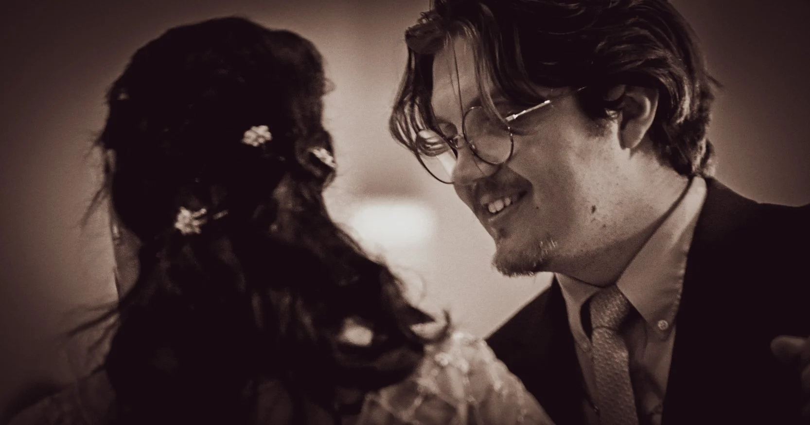 A man and woman smiling at each other closely, with the man wearing glasses and a suit and the woman with dark, curly hair.