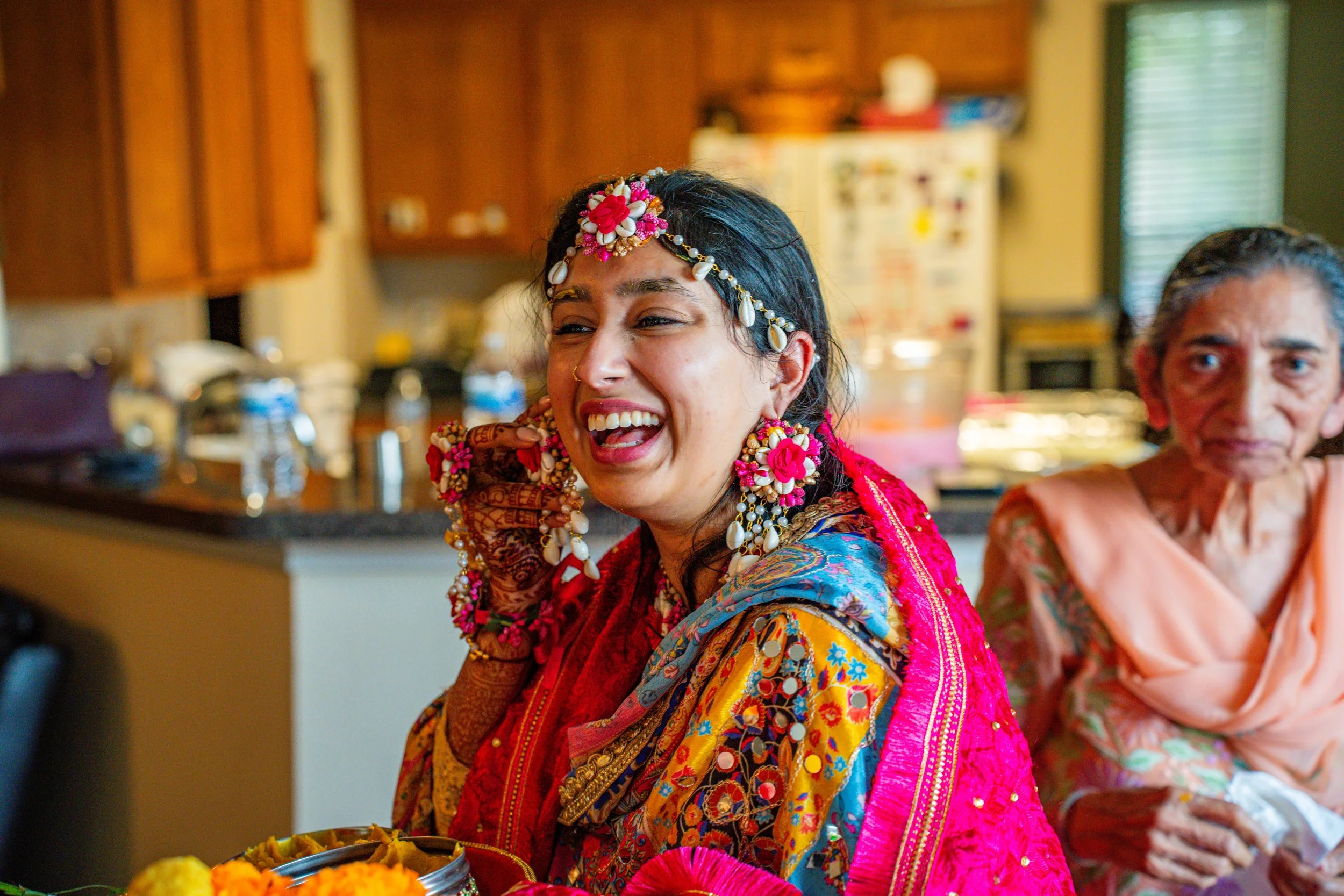 A woman dressed in traditional colorful Indian attire, adorned with jewelry and flowers, smiling and laughing, with an elderly woman sitting nearby in a kitchen setting.