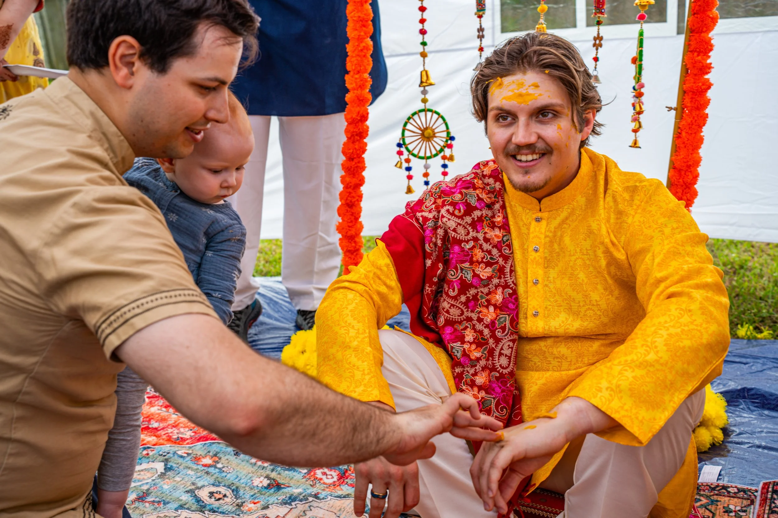 A man in traditional Indian attire with yellow and red clothing, sitting on the ground with turmeric paste on his forehead, during a cultural or religious ceremony. A man and a small child, possibly his family, are interacting with him. The backgroun
