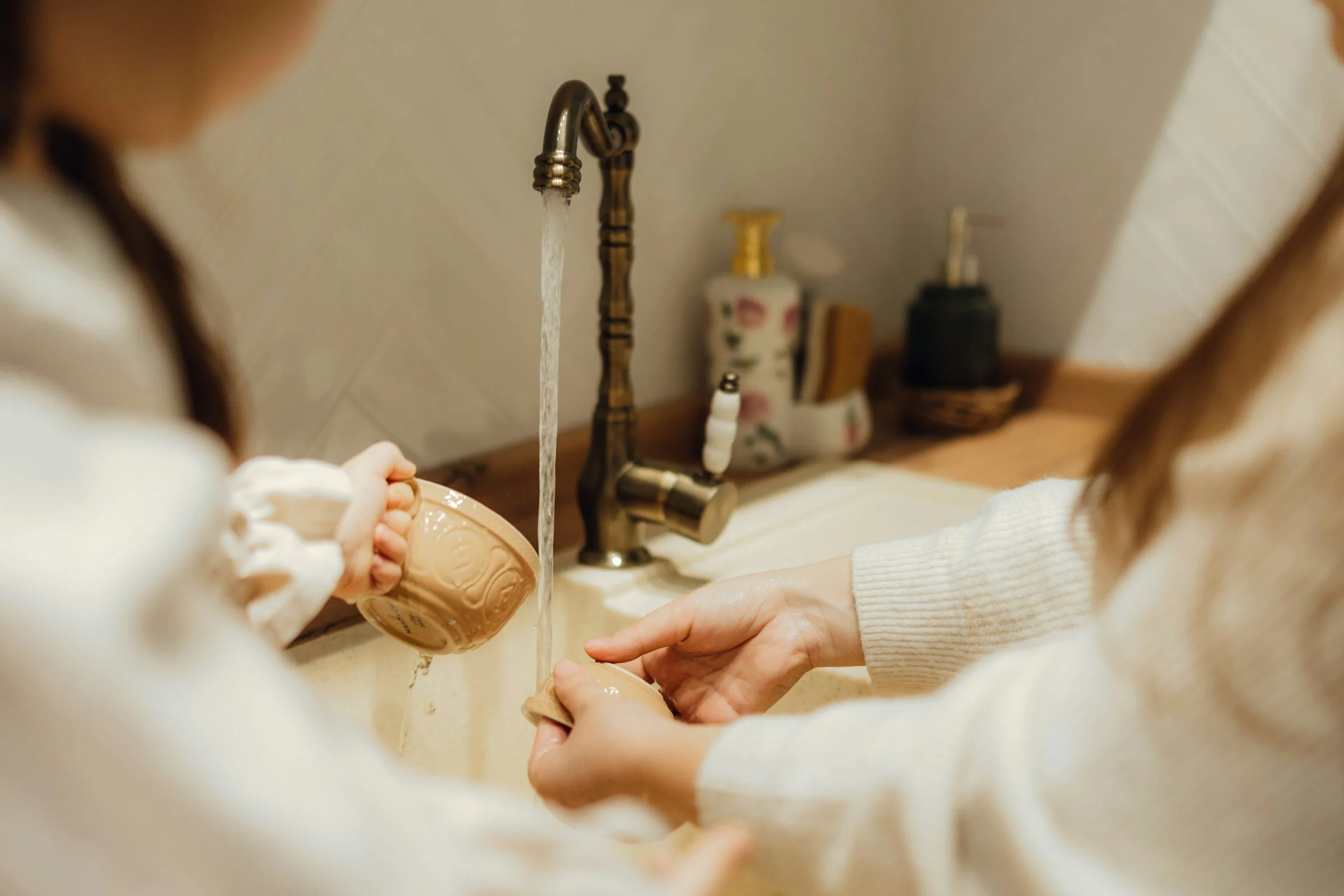 Two people rinsing a cup together under a kitchen tap