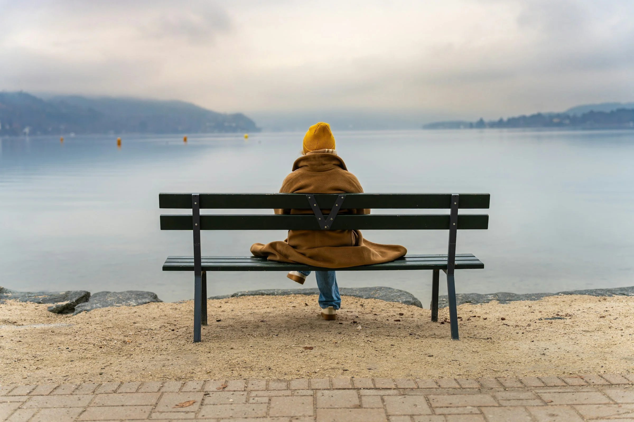A person in a yellow hat sitting alone on a bench by a calm lake
