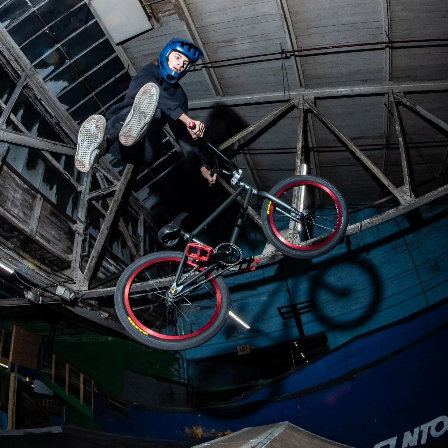 A person wearing a blue helmet performing a stunt on a black and red BMX bike inside a skate park or indoor riding facility.