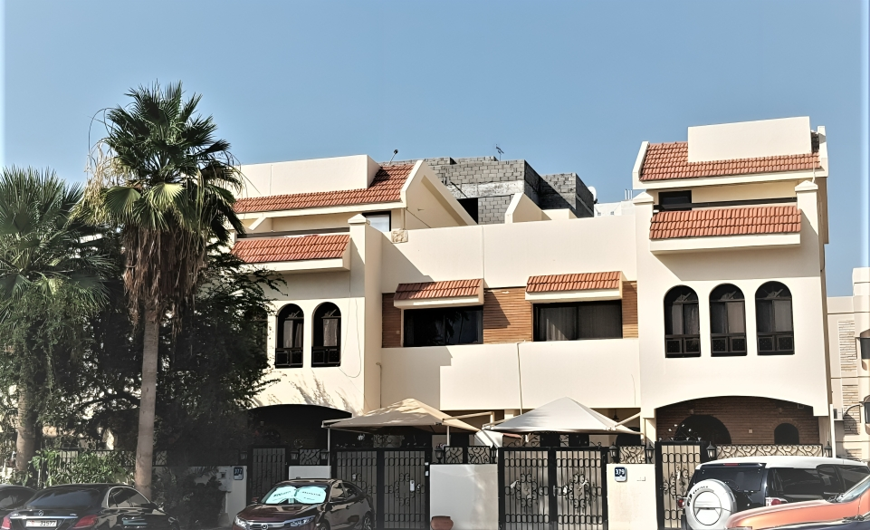 Large beige house with arched windows, red-tiled roof, palm trees, and cars parked outside.