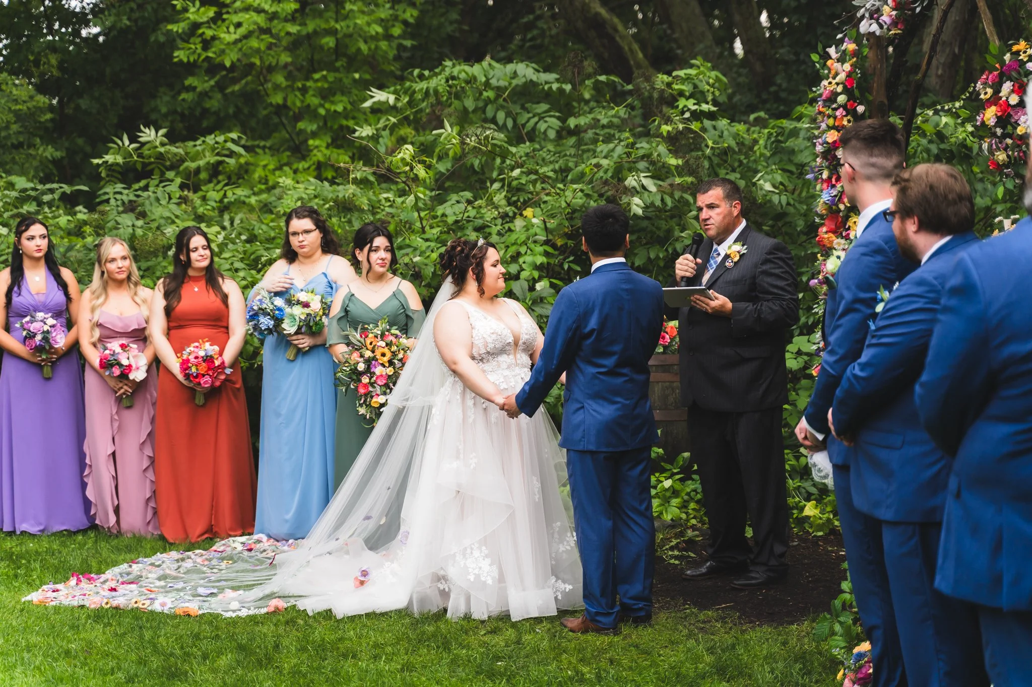 A wedding ceremony outdoors with a bride and groom holding hands and exchanging vows. Bridesmaids in colorful dresses are holding bouquets, while groomsmen and a officiant dress in blue suits, next to a floral arch.