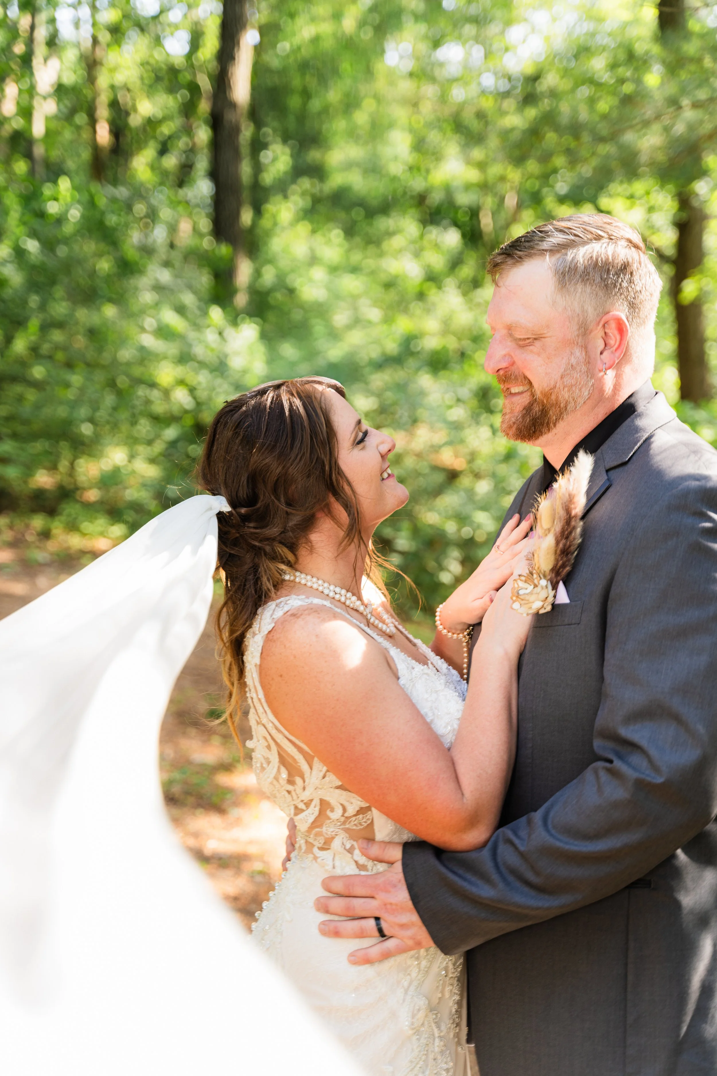 A bride and groom smile at each other in a wooded outdoor setting, with the bride in a lace wedding dress and the groom in a gray suit.
