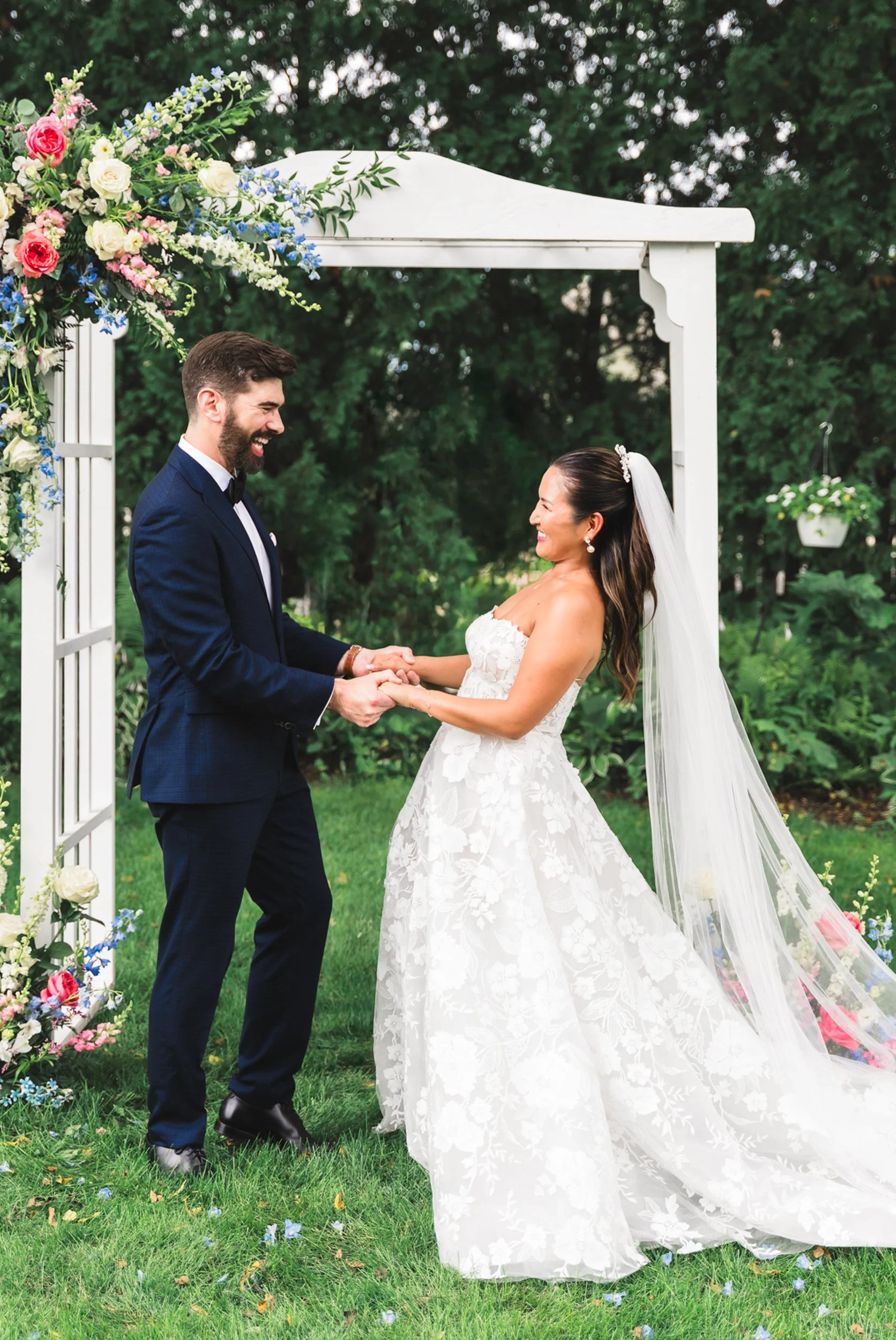 A bride and groom holding hands and smiling at each other during their wedding ceremony outdoors under a floral arch with green trees in the background.