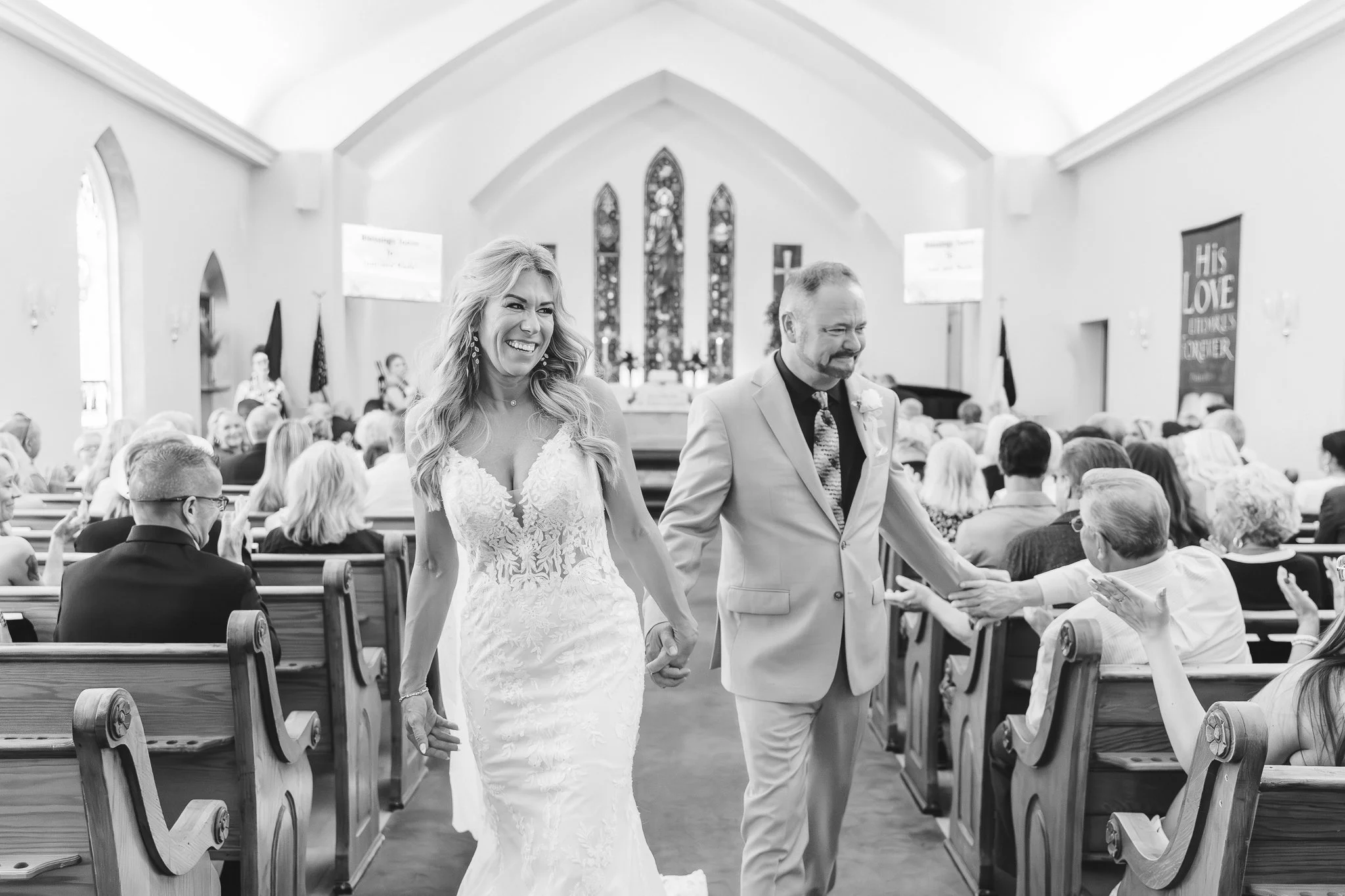 Wisconsin Wedding Photographer Church Ceremony | Black and white photo of Bride and groom walk down the isle as newly weds, holding hands and smiling,  groom gives handshake to friend and bride smile, small church congregation behind them