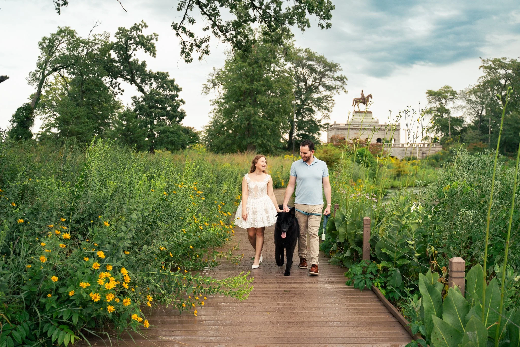 Lincoln Park Chicago Illinois Engagement Photographer | Bride in lace white dress holding hands with groom walking forward, black pet dog between them, Honeycomb in background | Midwest Wedding and Engagement Photography Sessions