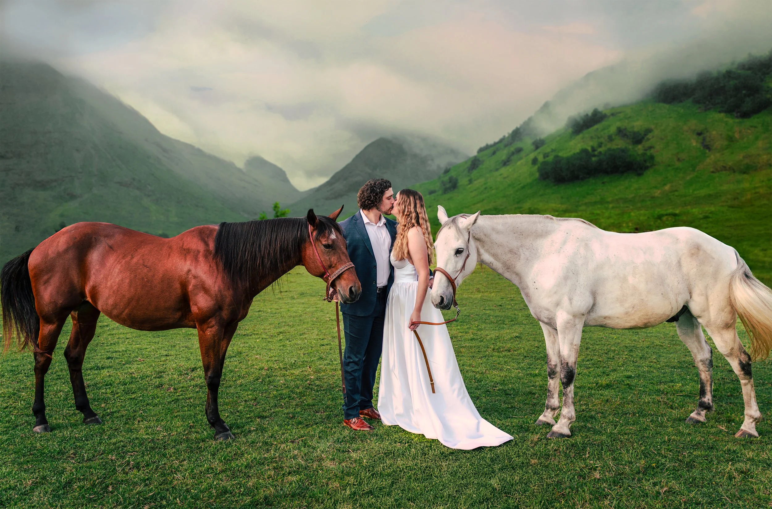 A couple dressed in wedding attire, holding horse leads, standing on a grassy field with two horses, one brown and one white, in front of green mountains and cloudy sky.