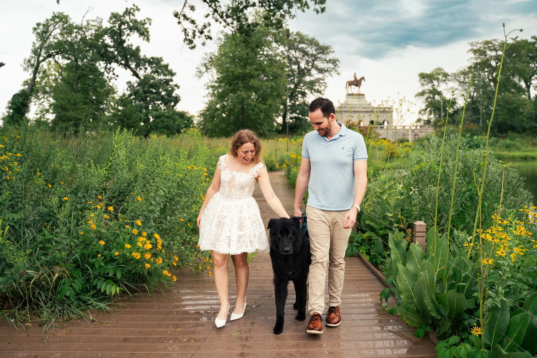 Lincoln Park Chicago Illinois Engagement Photographer | Bride in lace white dress holding hands with groom walking forward, black pet dog between them, Honeycomb in background | Midwest Wedding and Engagement Photography Sessions